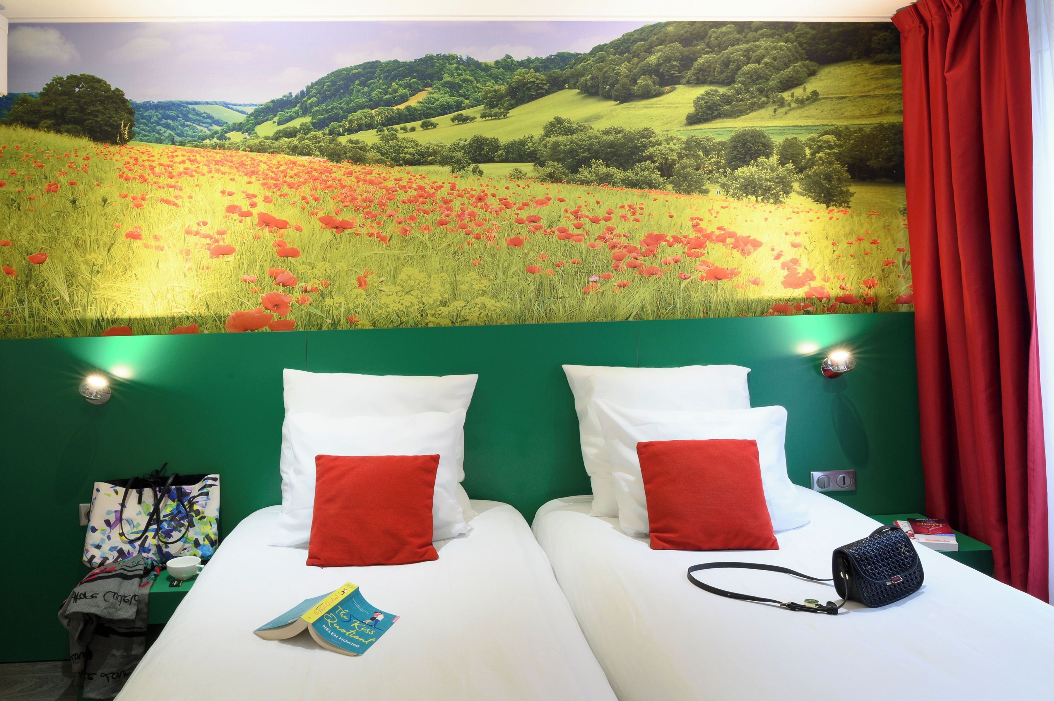 A hotel room with two beds, red and white pillows, a green headboard, a red curtain, and a landscape mural on the wall.