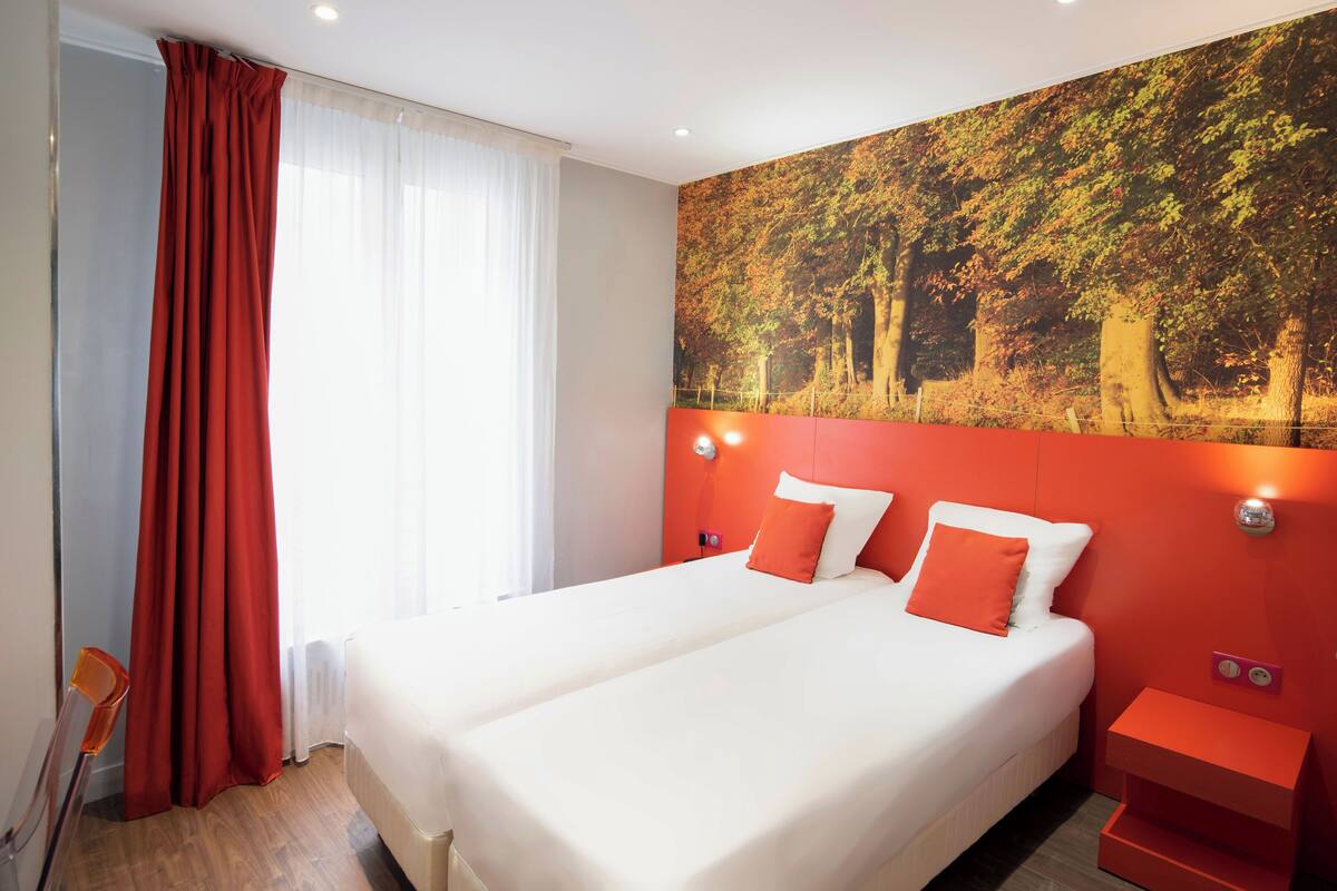 A double bed with white and orange bedding, a red accent wall, and a large forest-themed painting above the bed.