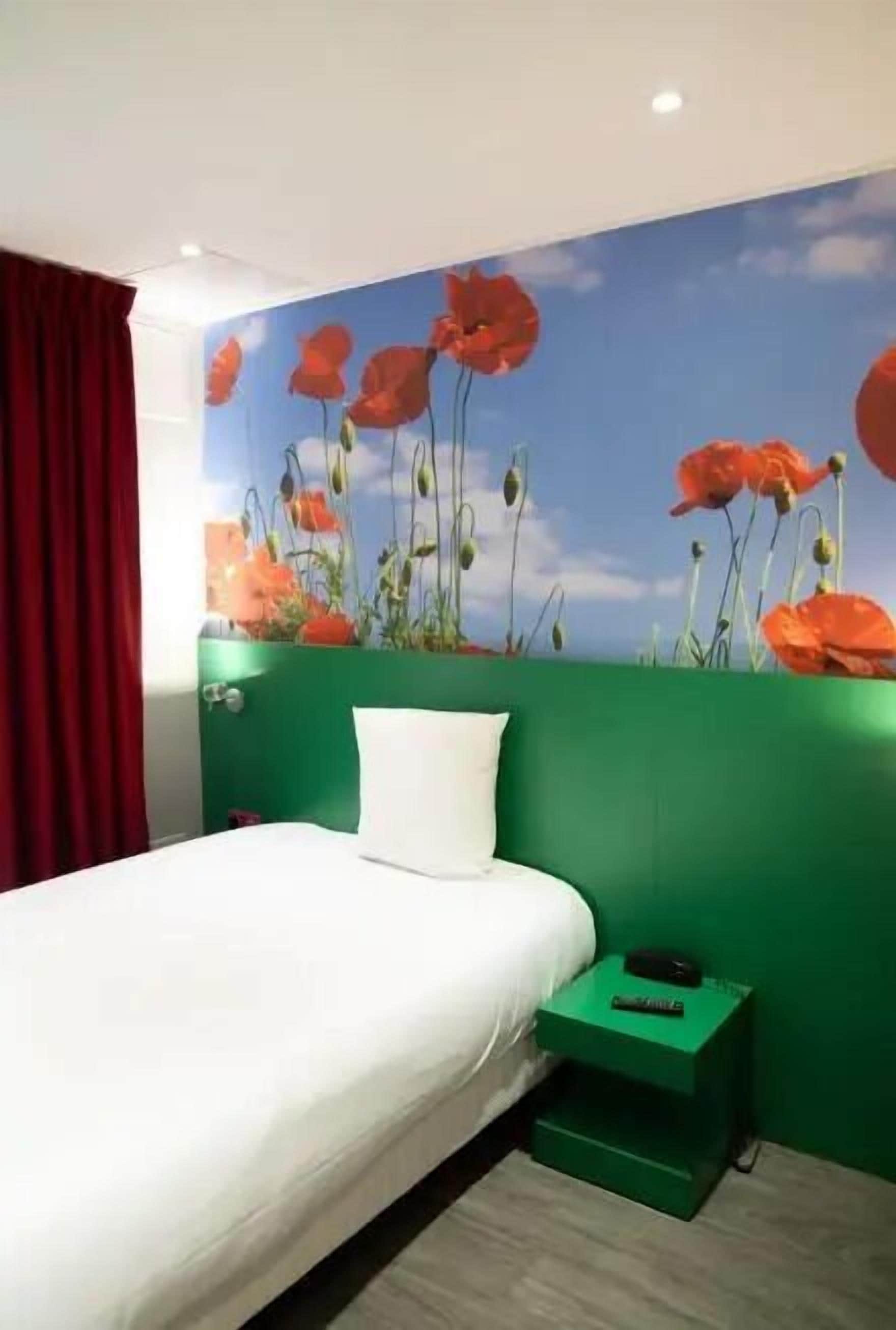 A hotel room with a bed, green walls, a red curtain, and a mural of red flowers on the wall.