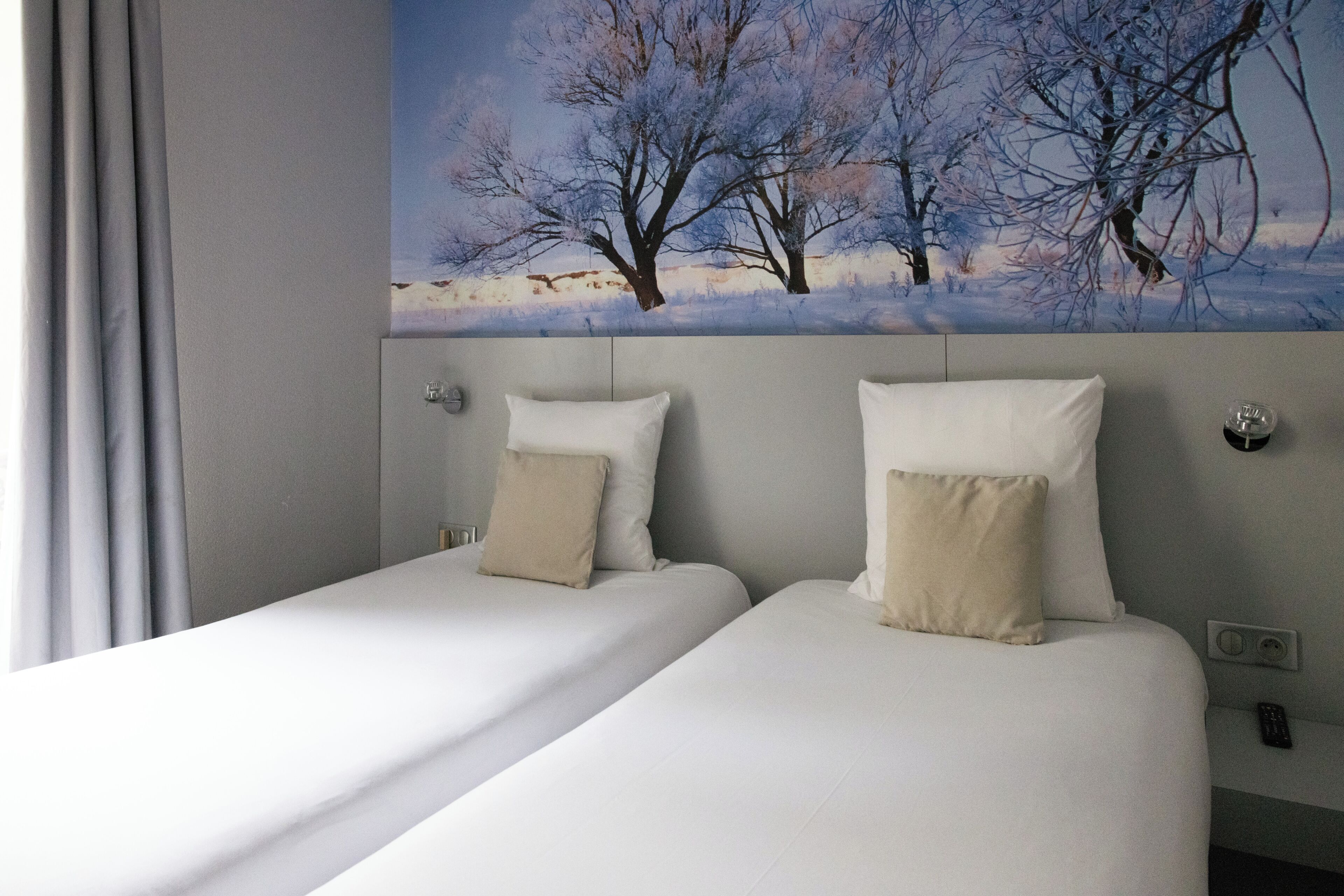 A hotel room with two beds, a painting of trees on the wall, and a window with curtains.