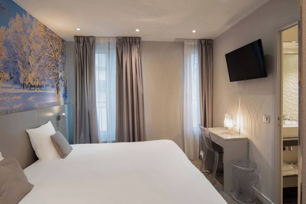 A hotel room with a large bed, a desk with a chair, a TV mounted on the wall, and a bathroom visible through an open door.