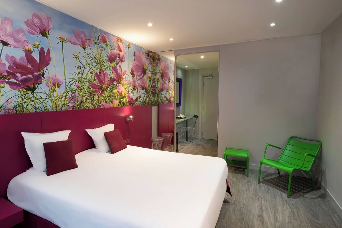 A modern hotel room with a large bed, a vibrant floral mural, a green bench, and a glass door leading to a dining area.