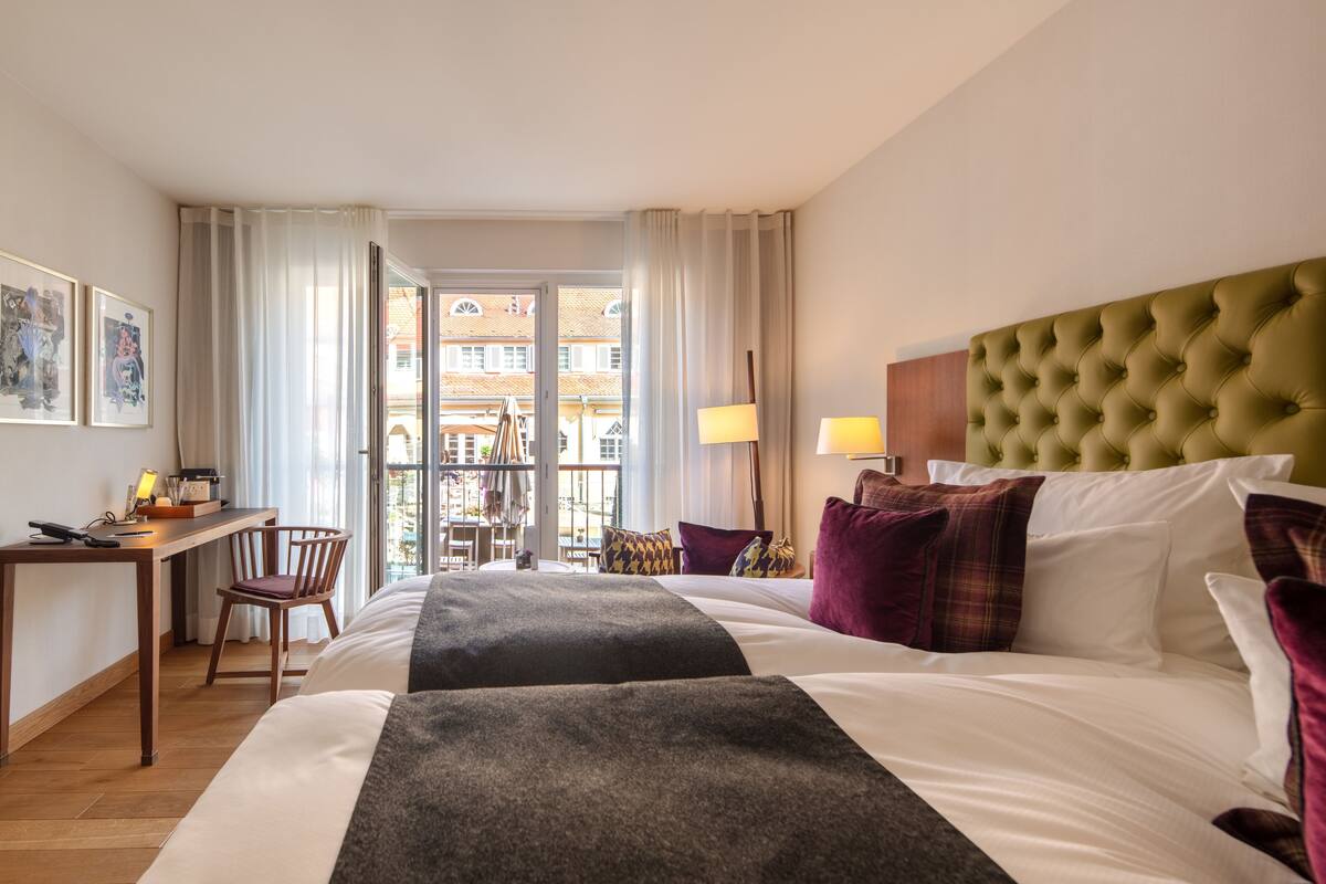 A hotel room with a bed, a desk, a chair, a lamp, and a view of the outside.