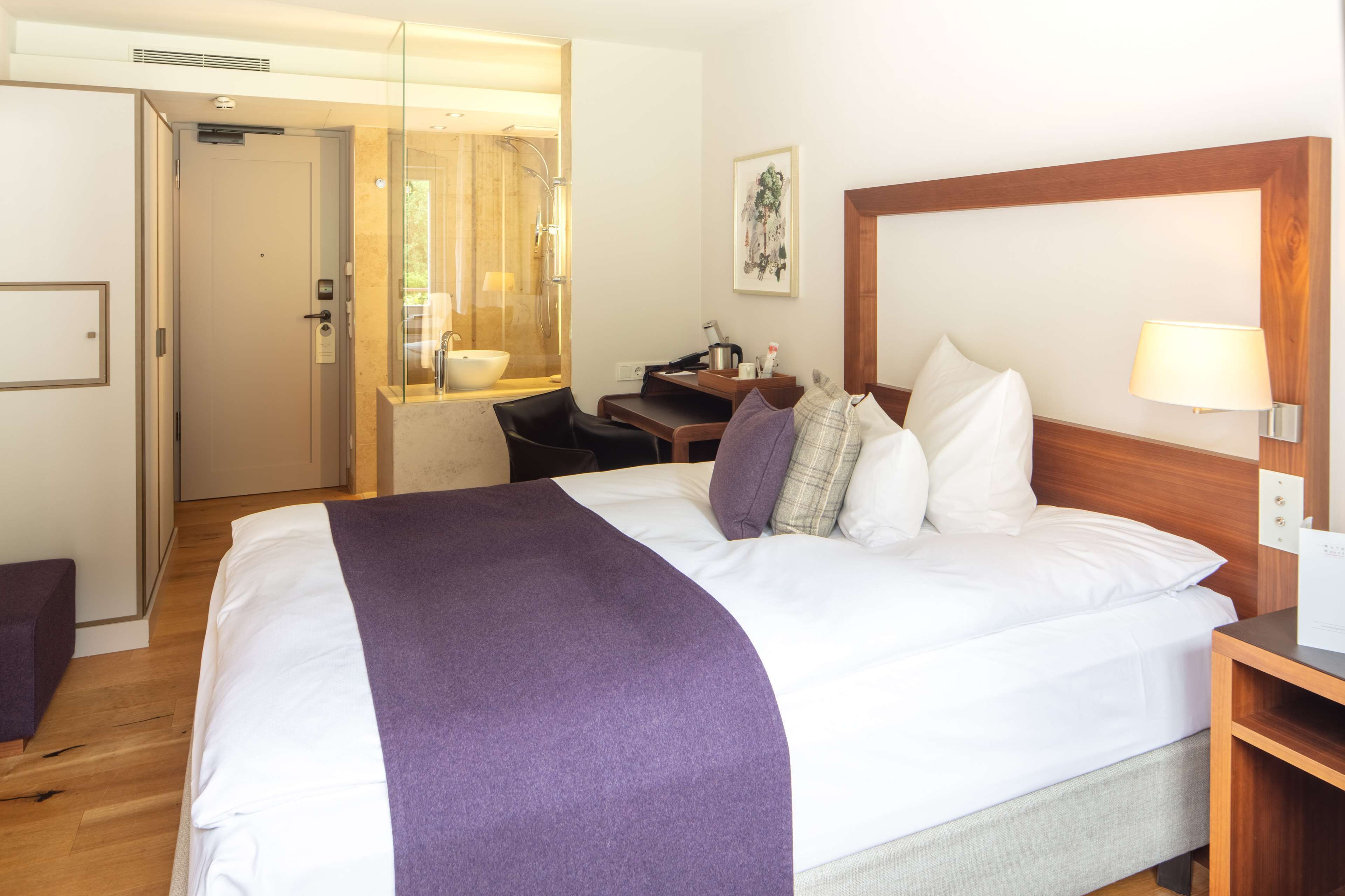 A hotel room with a bed, a desk, a chair, and a bathroom visible through a glass door.