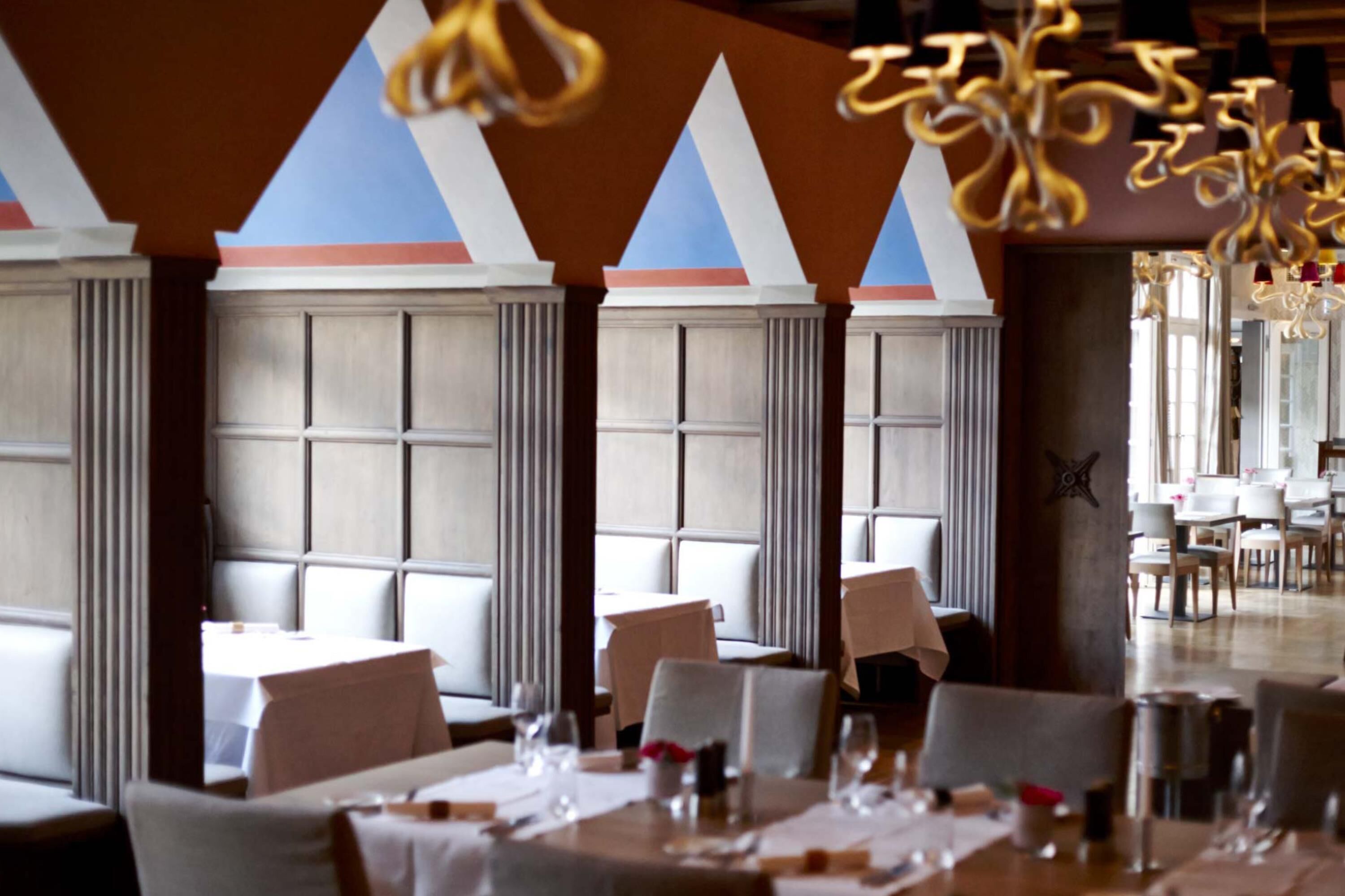 Un restaurante con techos altos, ventanas amplias y candelabros modernos. Las mesas están preparadas con manteles blancos y cristalería.