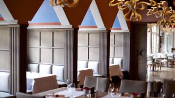 Un restaurante con techos altos, ventanas amplias y candelabros modernos. Las mesas están preparadas con manteles blancos y cristalería.