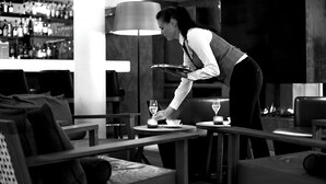 A bar area with a bartender setting a glass on a table. There are multiple tables and chairs, and a bar stocked with bottles.