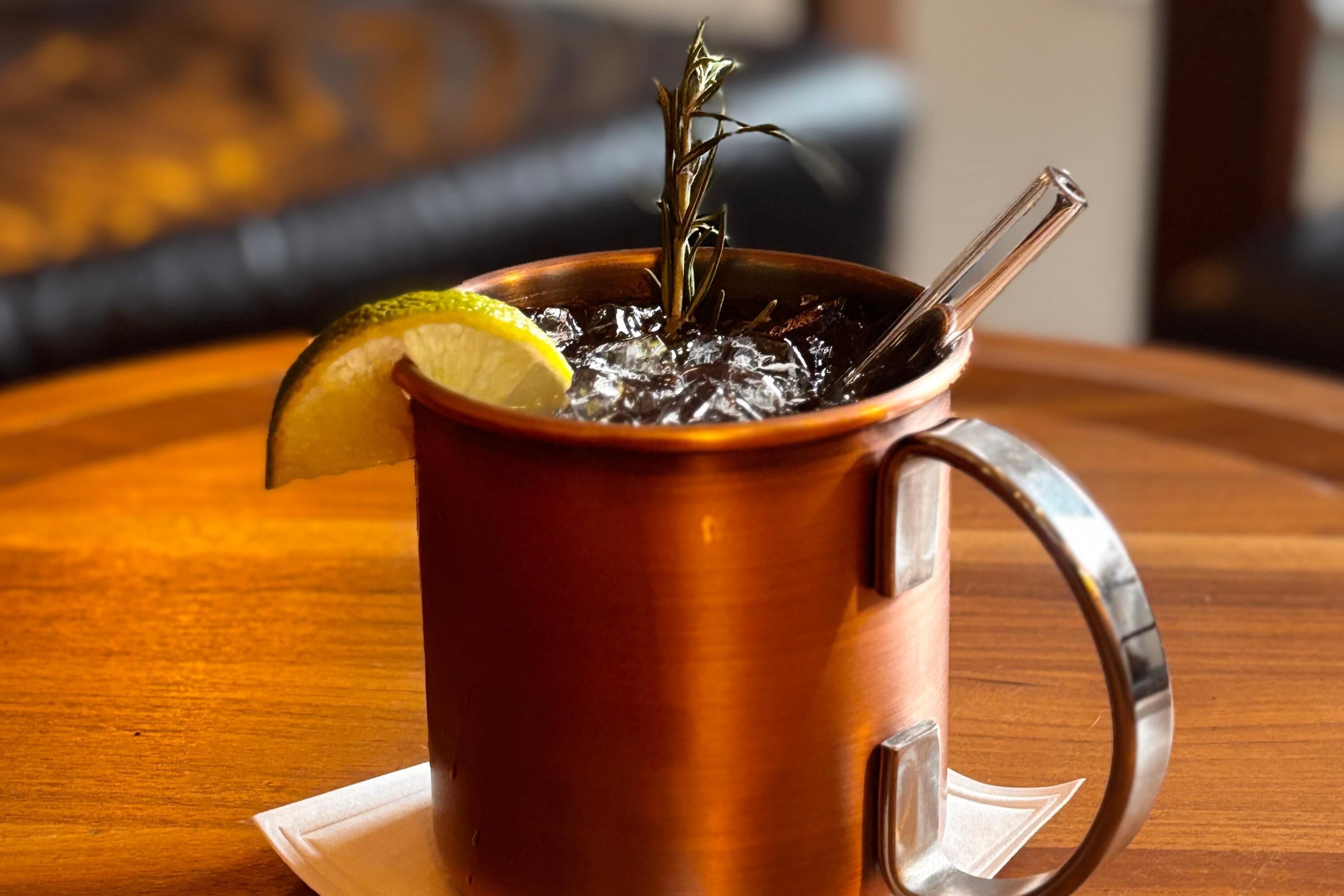 A copper mug with a lemon slice and a sprig of rosemary on a wooden table.