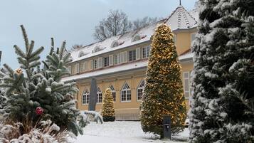 A building with a yellow facade and multiple windows, surrounded by snow-covered trees and a Christmas tree with lights.