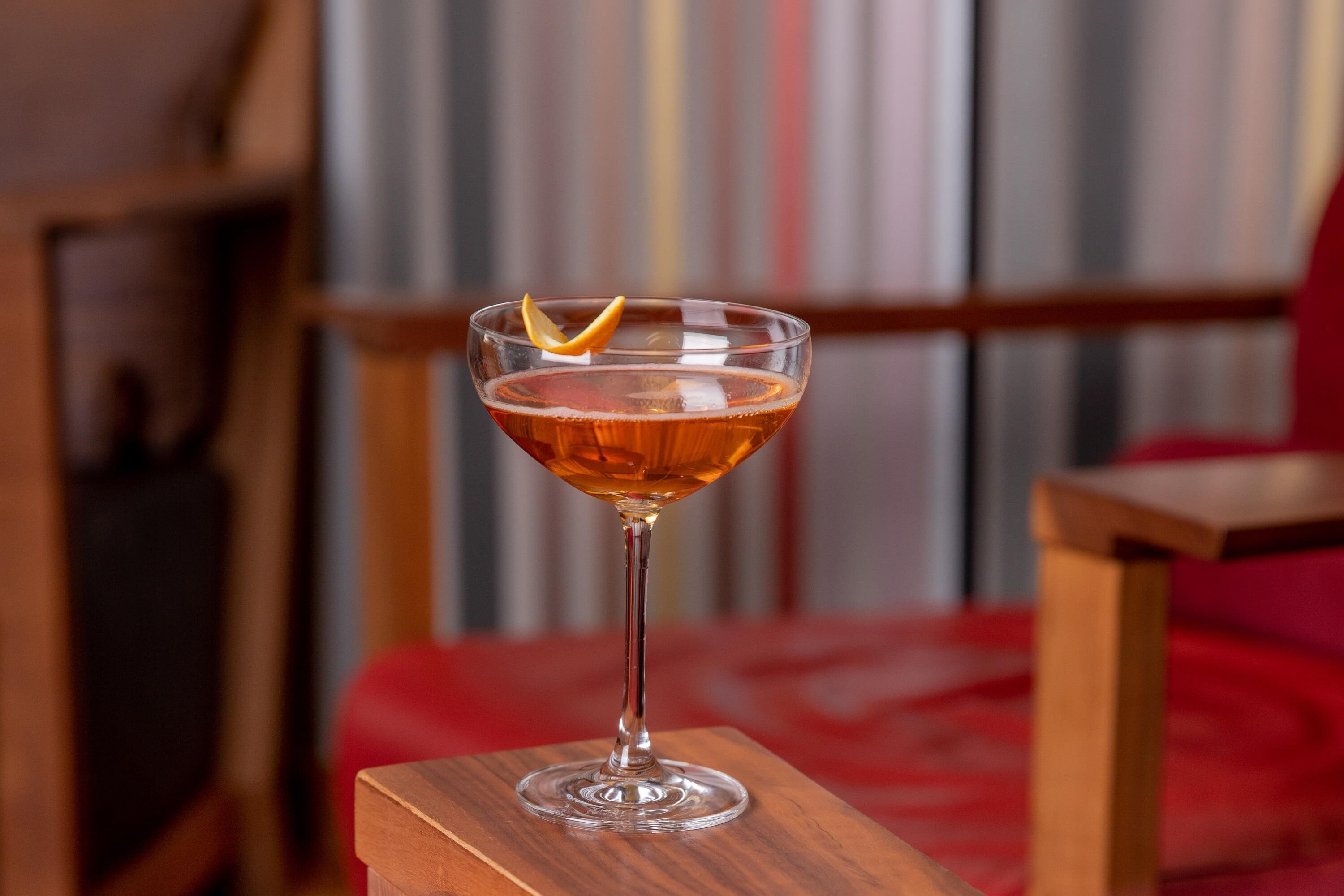 Ein Cocktail im Glas auf einem Holztisch, im Hintergrund ein rotes Kissen.