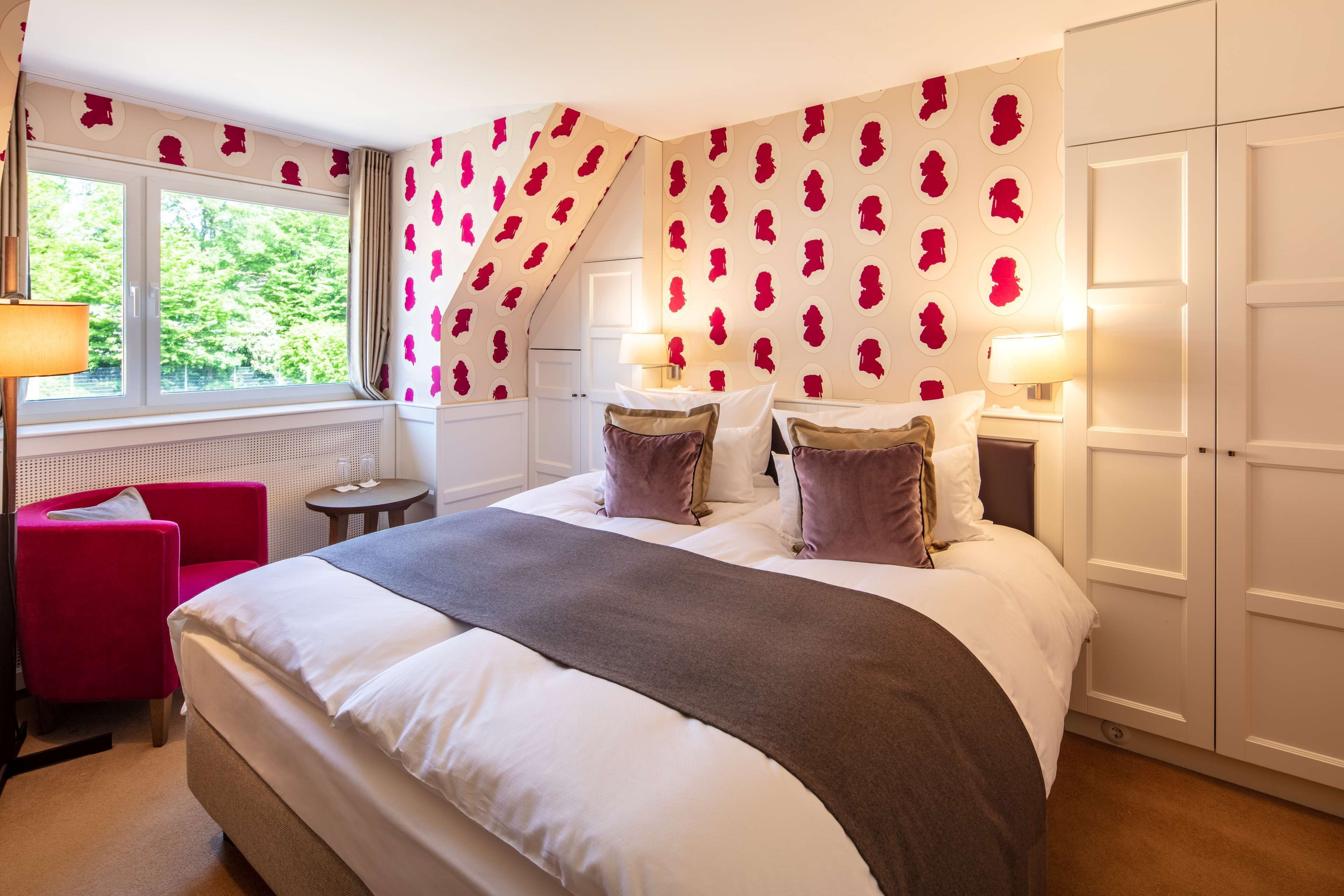 Habitación de hotel con una cama grande, una silla roja, una mesita pequeña y una ventana con cortinas.