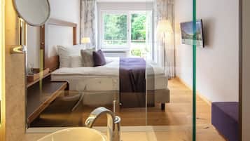 A hotel room with a bed, a bathroom with a bathtub, a mirror, and a window with a view of greenery.