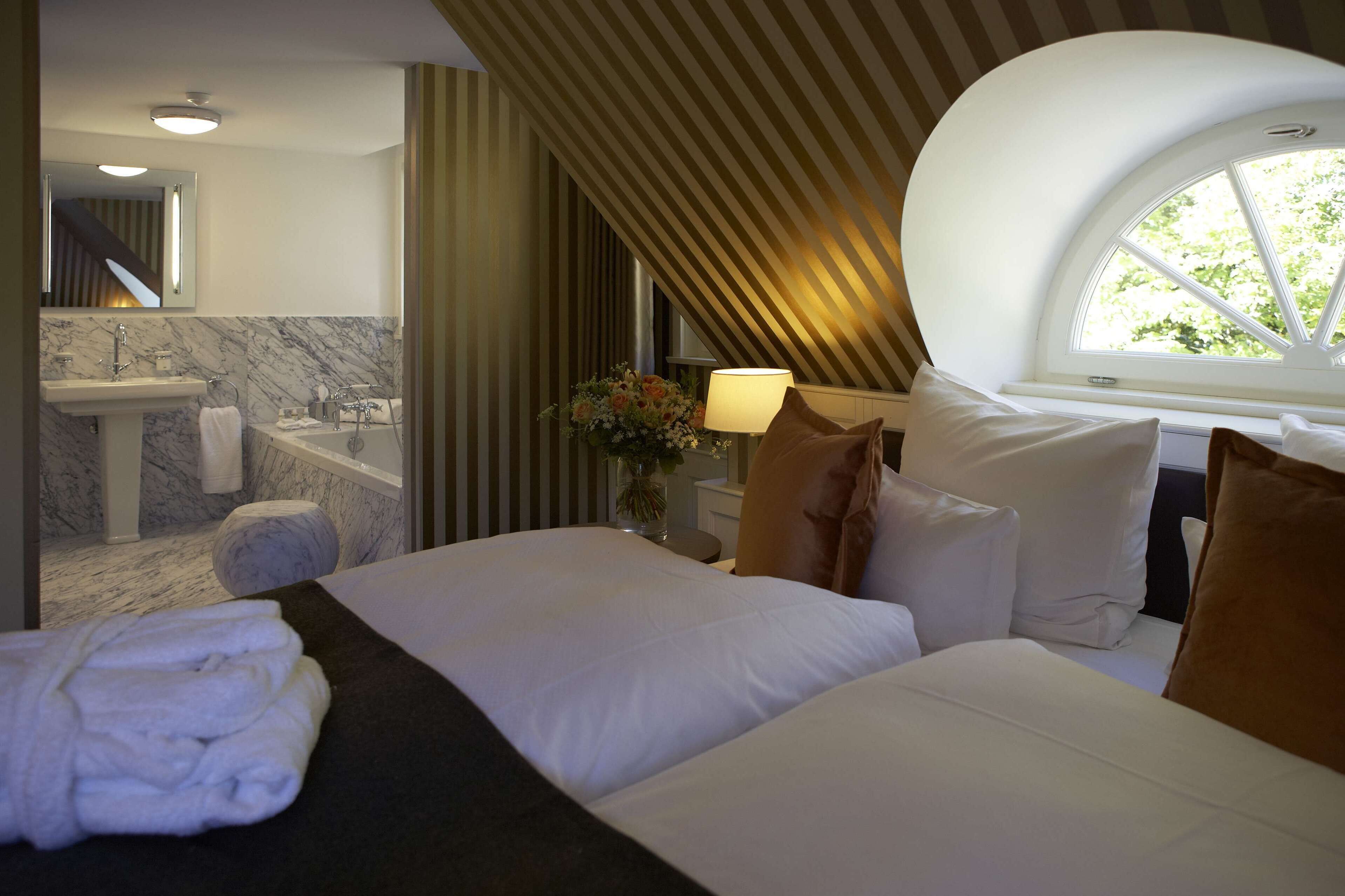 Habitación de hotel con cama, baño con lavamanos y bañera, ventana con vistas y una lámpara.