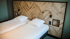 Un lit bien fait, avec du linge de lit blanc et deux oreillers, une tête de lit en bois à motifs chevrons, et deux tables de chevet fixées au mur, chacune avec une lampe.