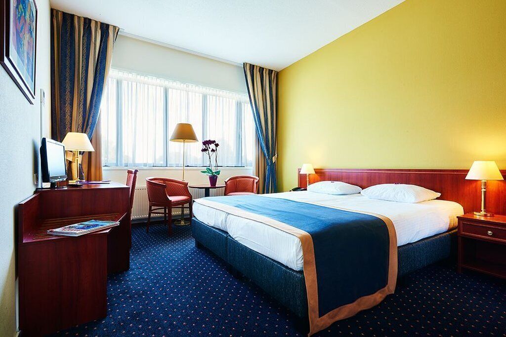 A hotel room with a bed, desk, chair, lamps, and a window with curtains.