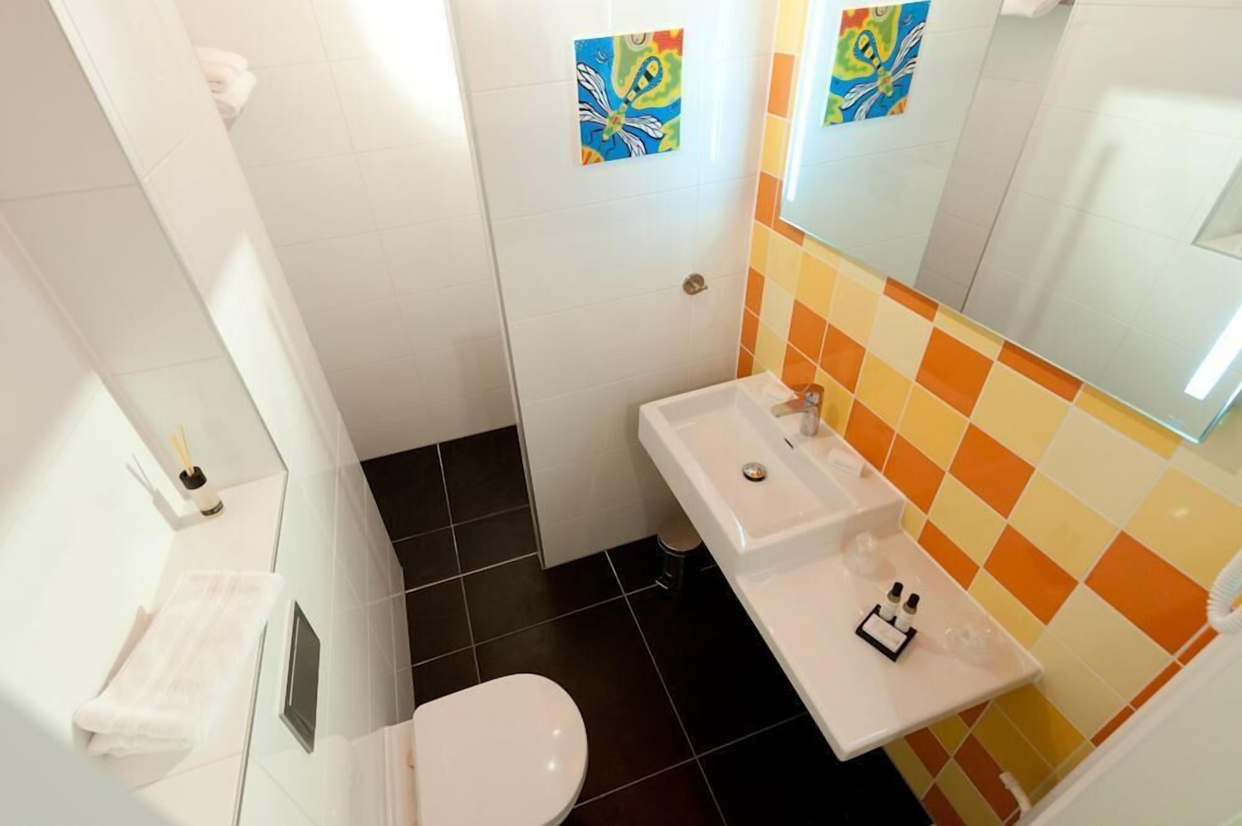 Une salle de bain avec des toilettes blanches, un lavabo et un miroir. Les murs sont recouverts de carreaux colorés et il y a une petite poubelle.
