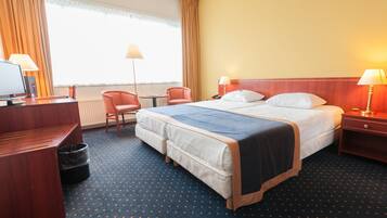 Ein Hotelzimmer mit einem großen Bett, zwei Nachttischen mit Lampen, einem Schreibtisch mit Computer, einem Stuhl und einem Fenster mit Vorhängen.