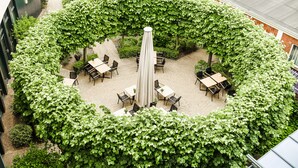 Un espace patio extérieur doté d’une table centrale ombragée par un parasol, entourée de chaises, et bordé d’une haie verdoyante et luxuriante.