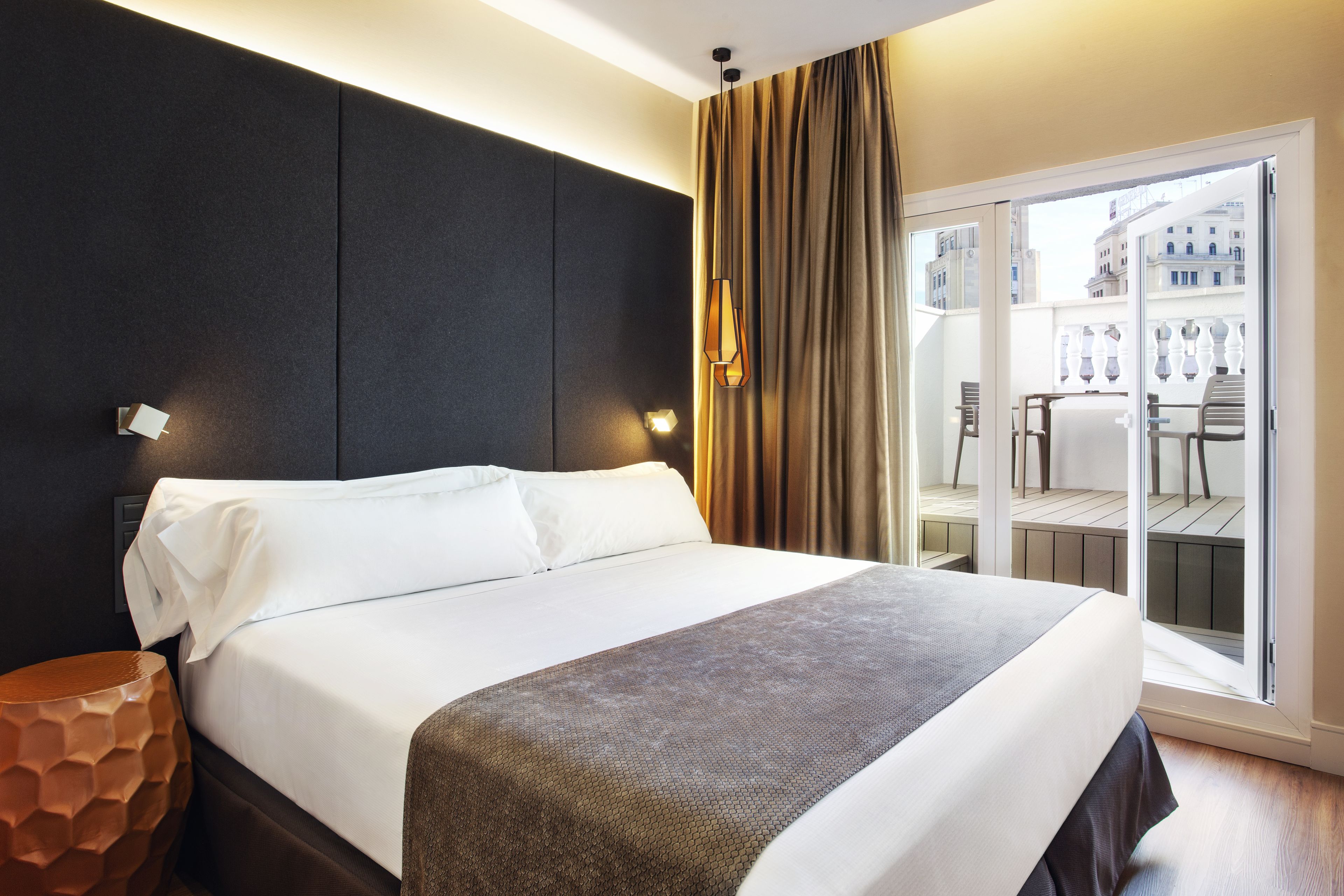 Una camera d'albergo moderna con un letto grande, un comodino, una lampada originale dalla texture particolare e vista sulla città tramite una porta scorrevole in vetro.