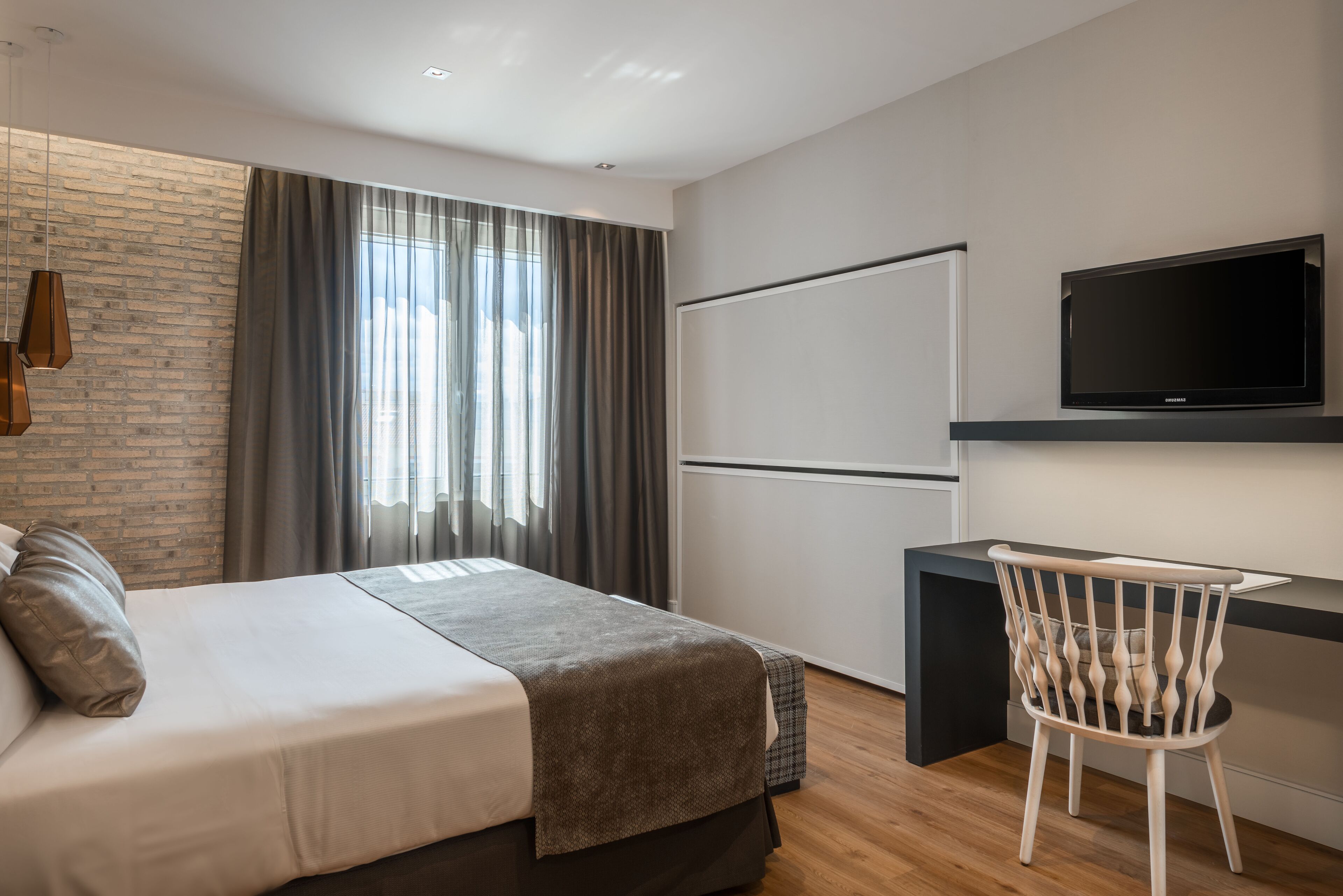 Una habitación de hotel moderna con una cama grande, un escritorio con una silla, un televisor de pantalla plana y un ventanal con cortinas.