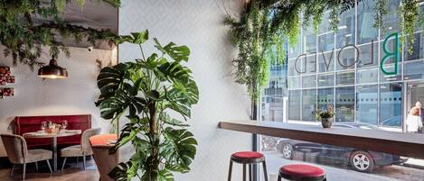 An interior with a large potted plant, bar stools, a red cushioned bench, and a view of the street through a window.