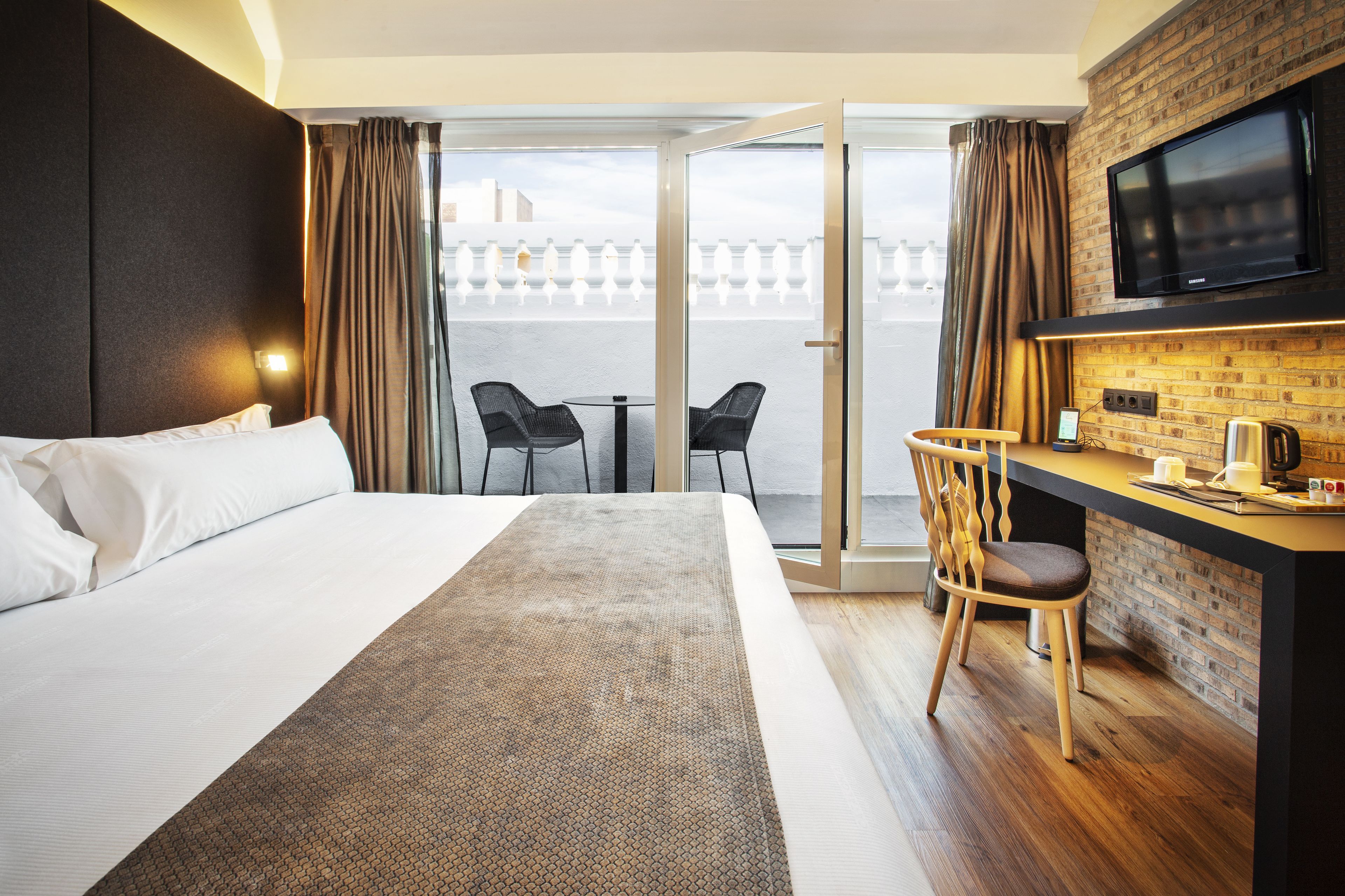 A hotel room with a bed, a desk with a chair, a TV, and a balcony with chairs.