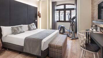 Ein modernes Hotelzimmer mit einem großen Bett, einem Schreibtisch, einem Sessel und einem Fitnessfahrrad, sowie einem Fenster mit Vorhängen.