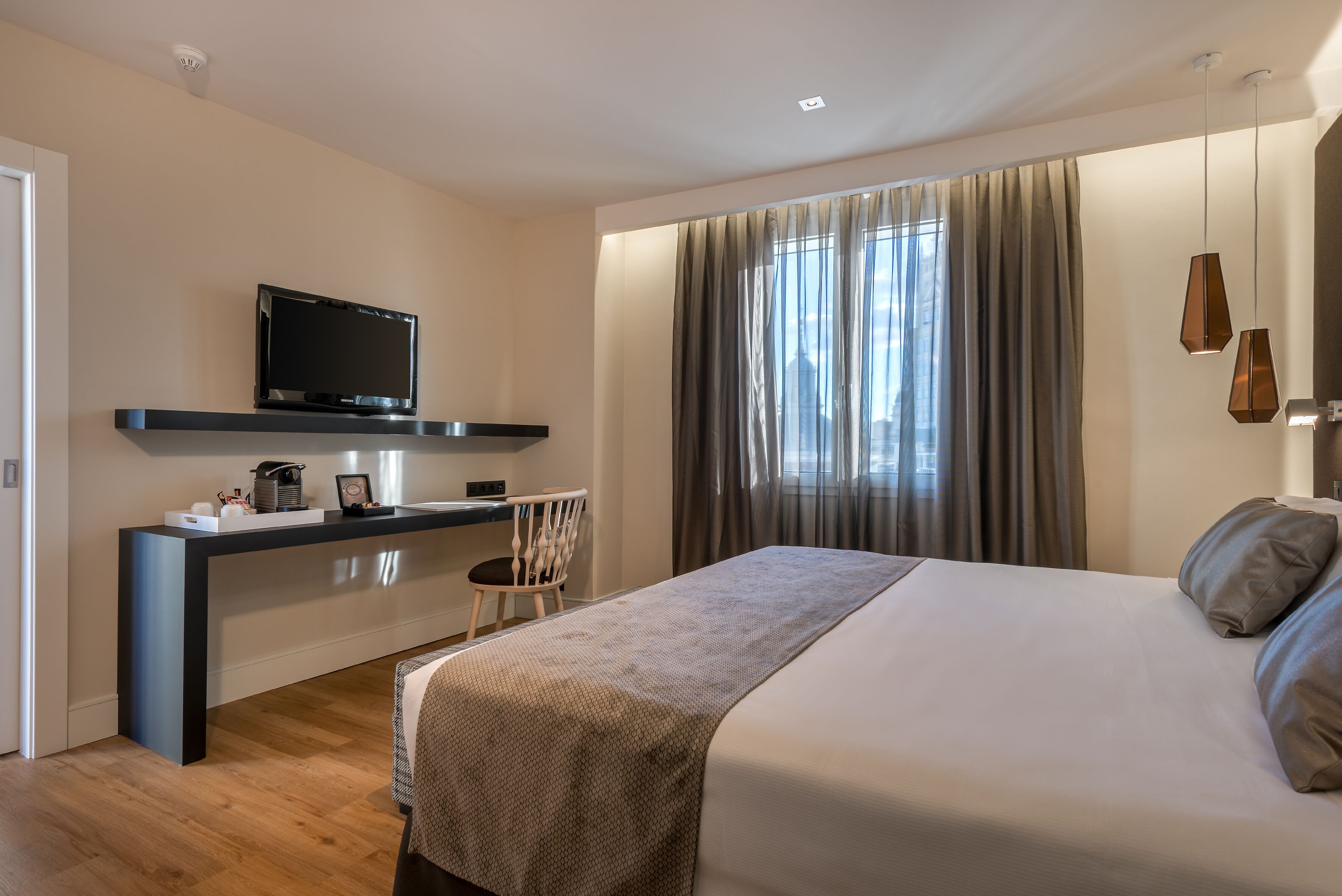 A modern hotel room with a large bed, a desk with a flat-screen TV, and a view of the city through large windows.