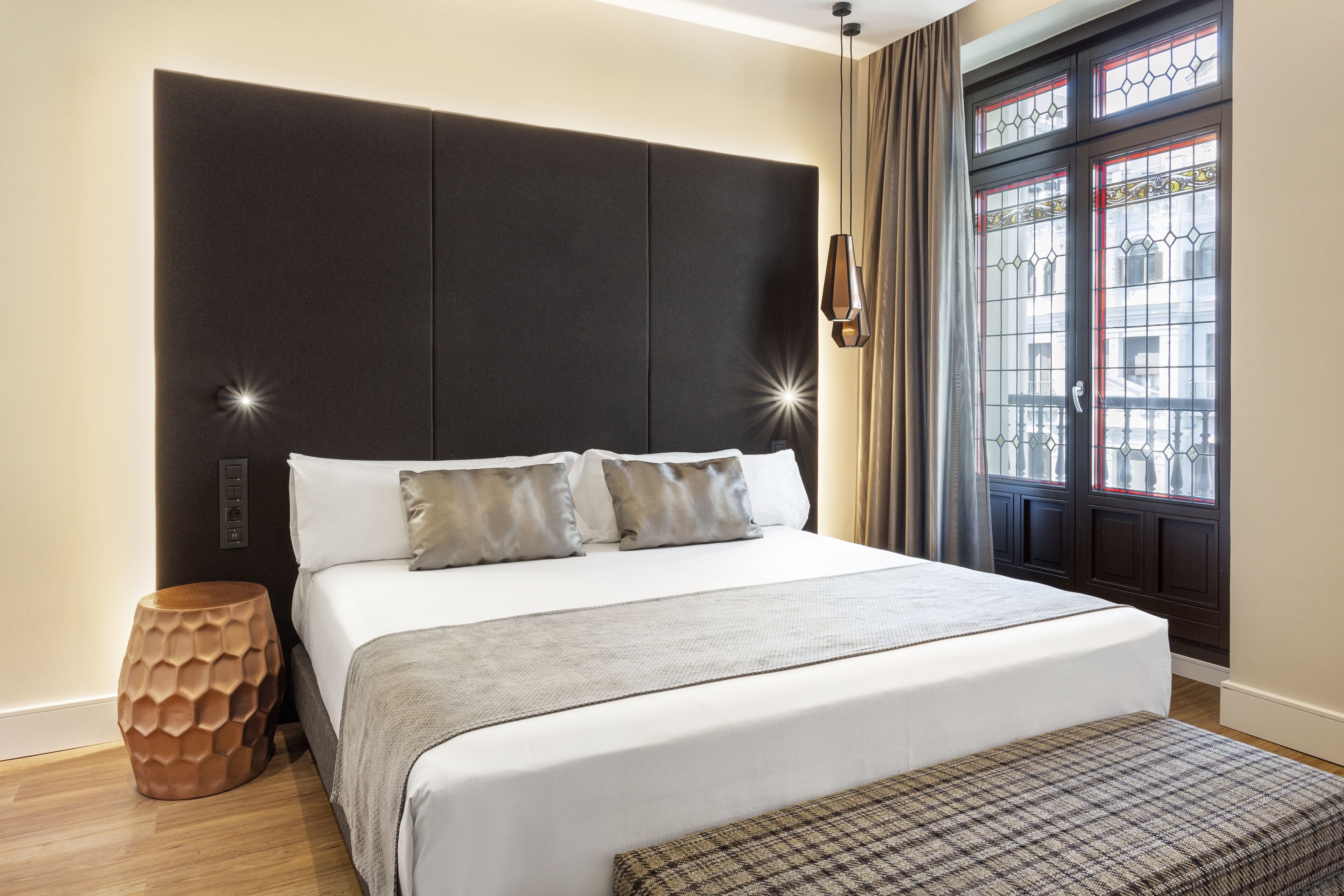 Een moderne hotelkamer met een groot bed, een houten nachtkastje en uitzicht op de stad door een raam met decoratief glas.