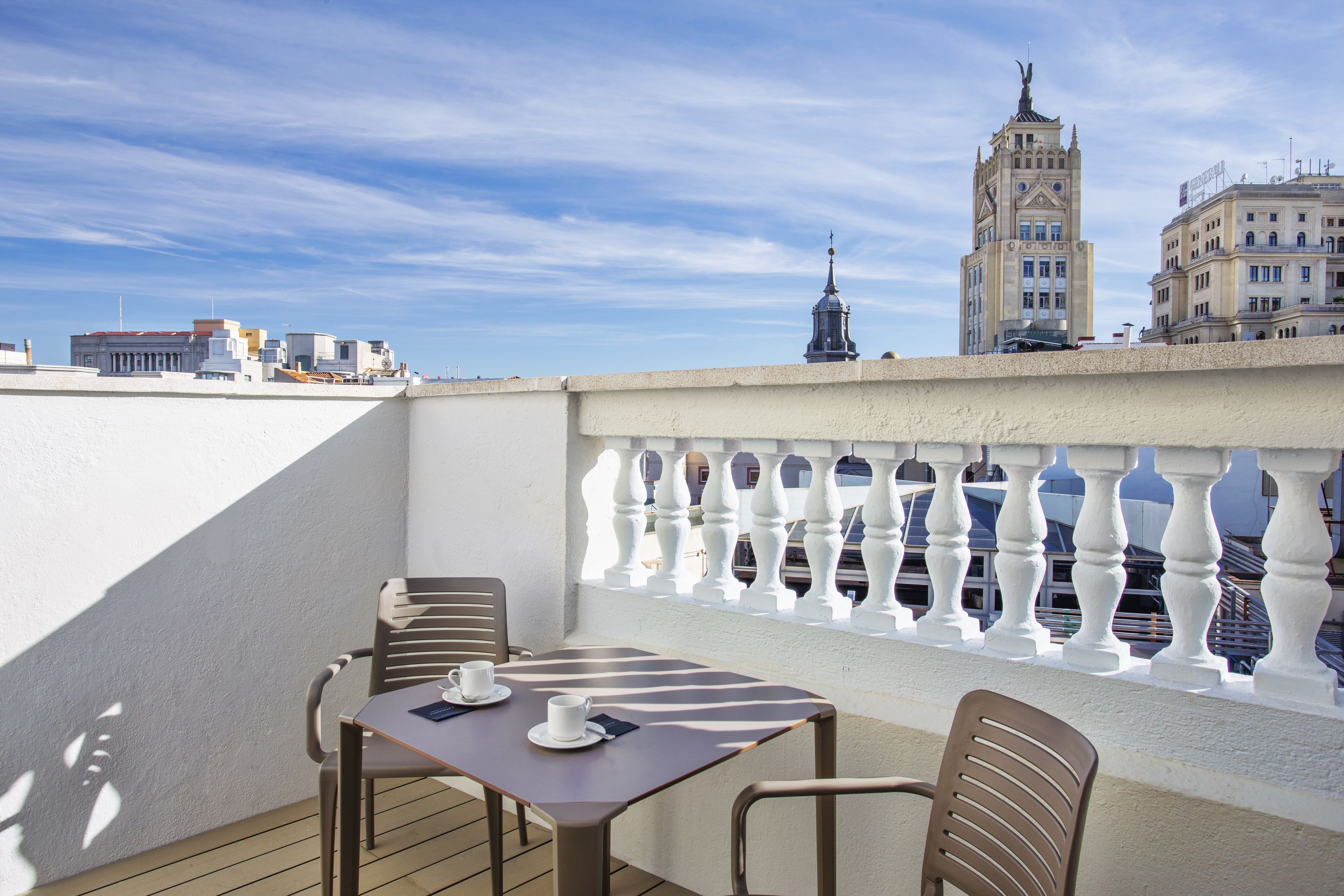 Ein Balkon mit Tisch und Stühlen, einem weißen Geländer und Blick auf Gebäude sowie einen Turm im Hintergrund.
