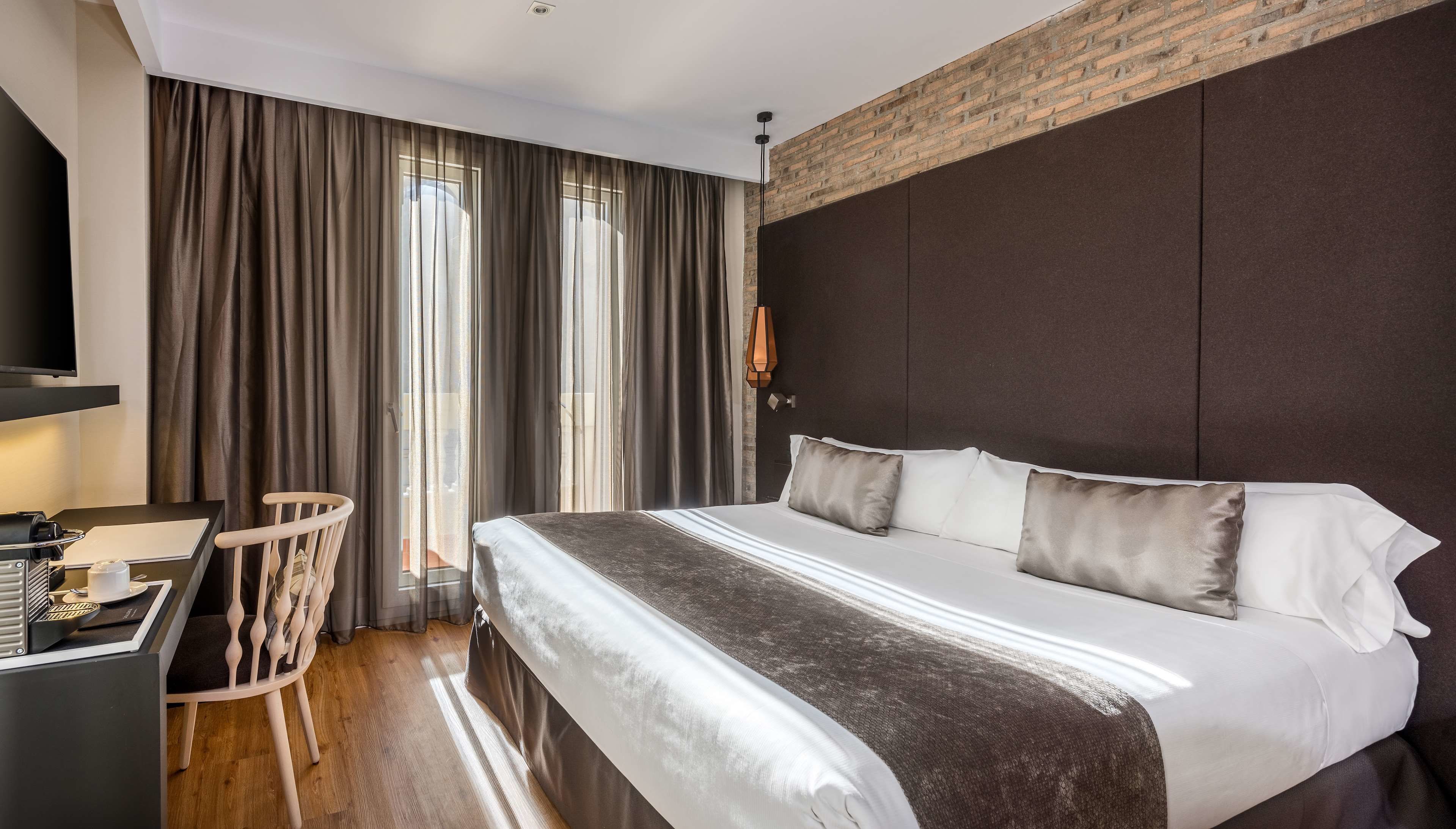 Una camera d'albergo moderna con un letto grande, una scrivania con una sedia e vista sulla città attraverso ampie finestre.