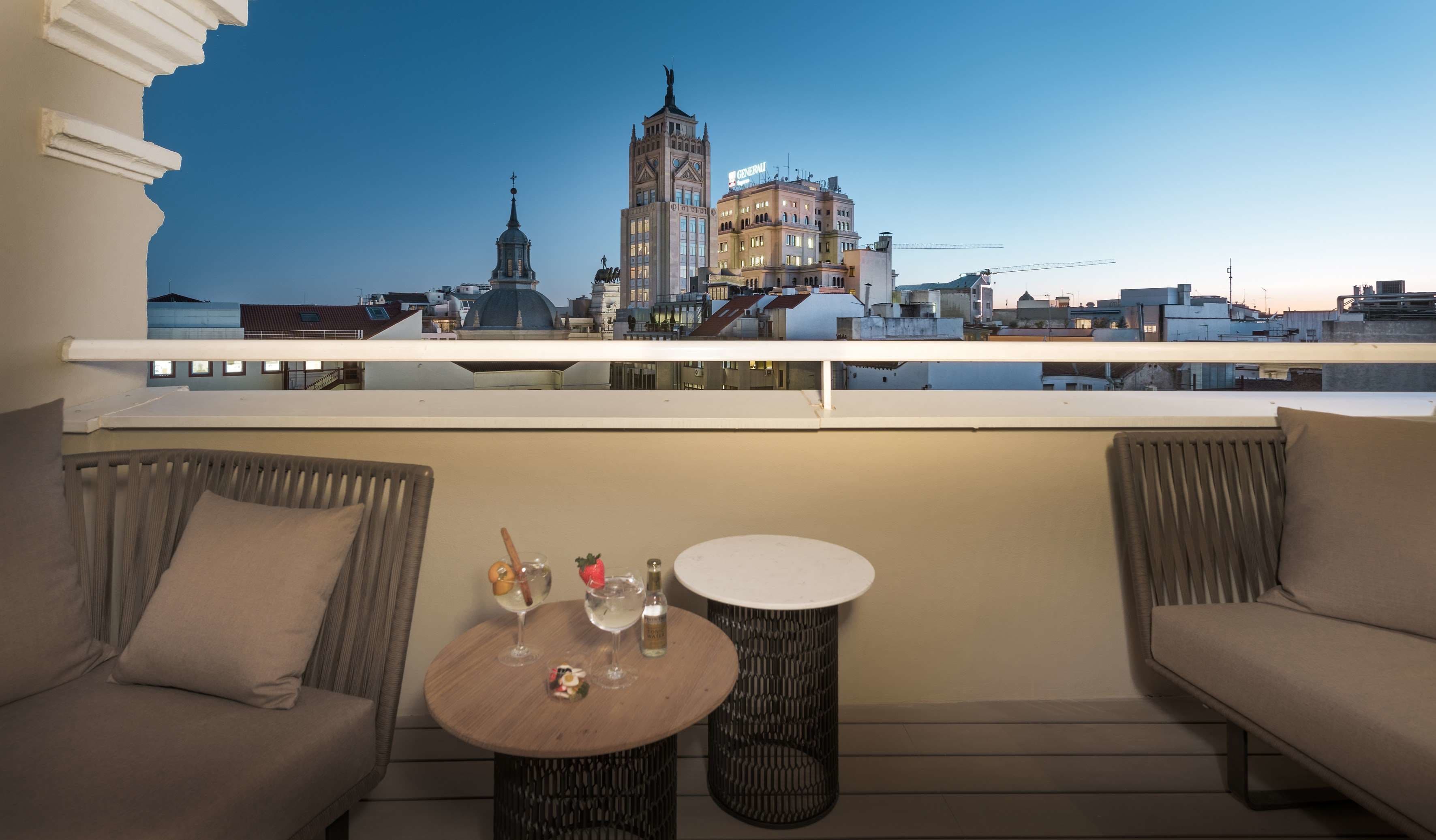 Un balcon avec vue sur un paysage urbain, comprenant une tour imposante et un bâtiment sur lequel on peut lire l’inscription « GENERATOR ». On y trouve deux tables rondes, dont l’une porte une bouteille et un verre, ainsi qu’un petit objet décoratif.