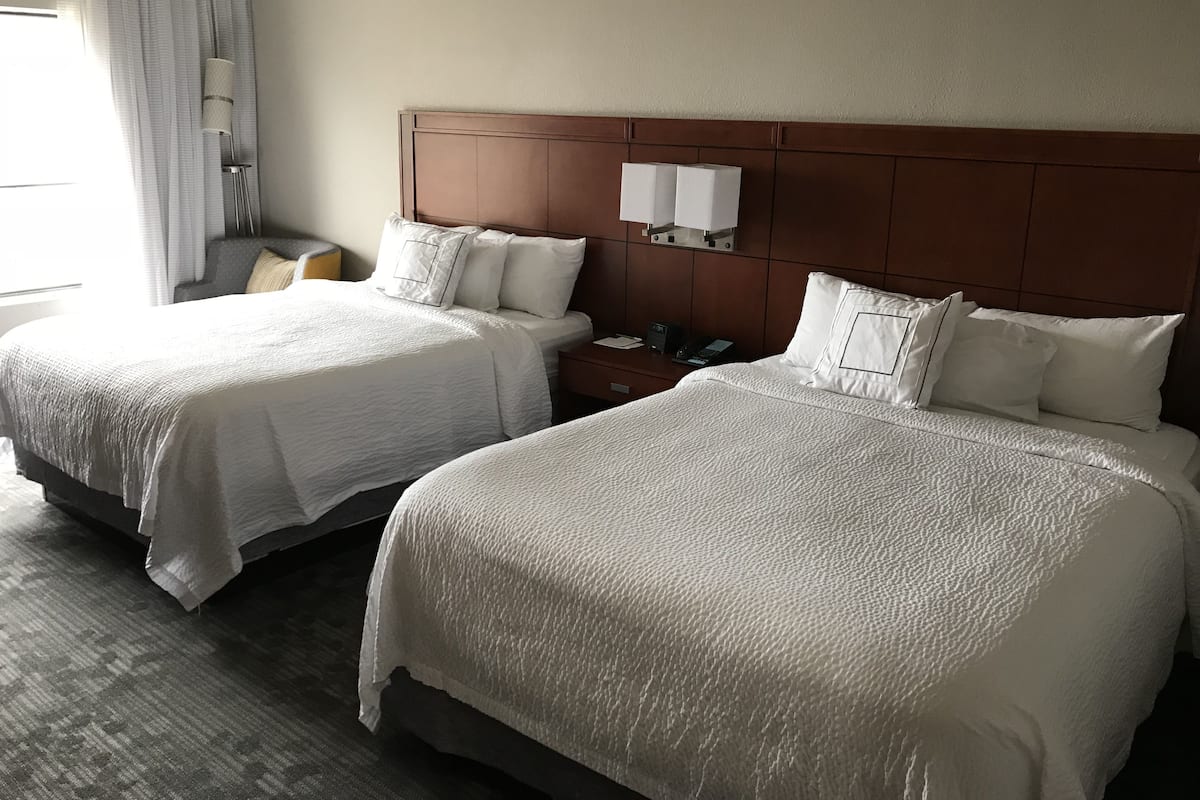 Two beds with white bedding, a wooden headboard, a bedside table with a lamp, and a window with curtains.