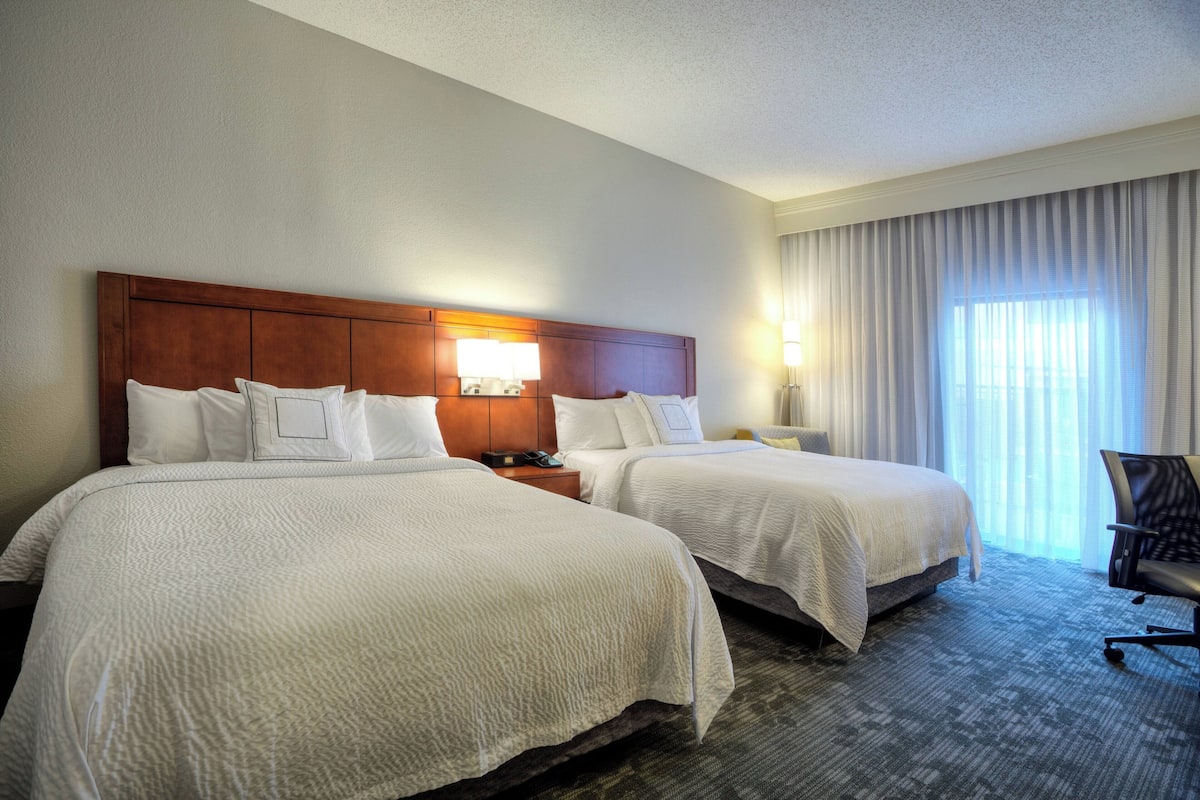 A hotel room with two beds, a wooden headboard, a nightstand, a lamp, and a view of the outdoors through large windows.