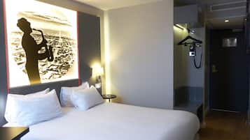 Habitación de hotel con cama, almohadas, mesita de noche, lámpara, televisor de pared mostrando una vista urbana y zona de ducha con toallero.