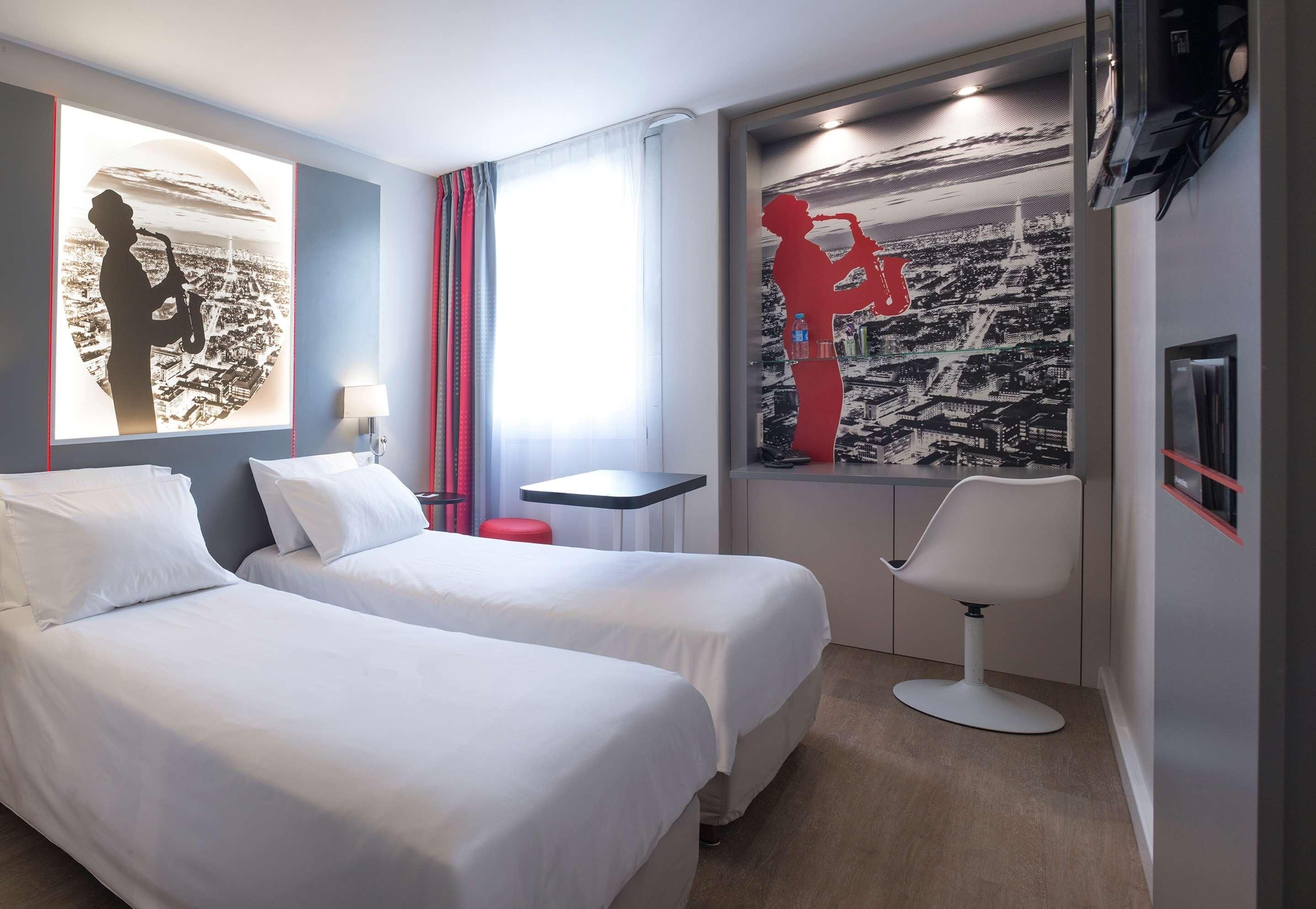 Une chambre d’hôtel avec deux lits, un bureau, une chaise et des œuvres d’art accrochées aux murs.