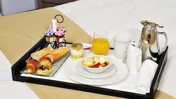 A breakfast tray with croissants, a bowl of fruit, a glass of orange juice, a coffee pot, and condiments on a bed.