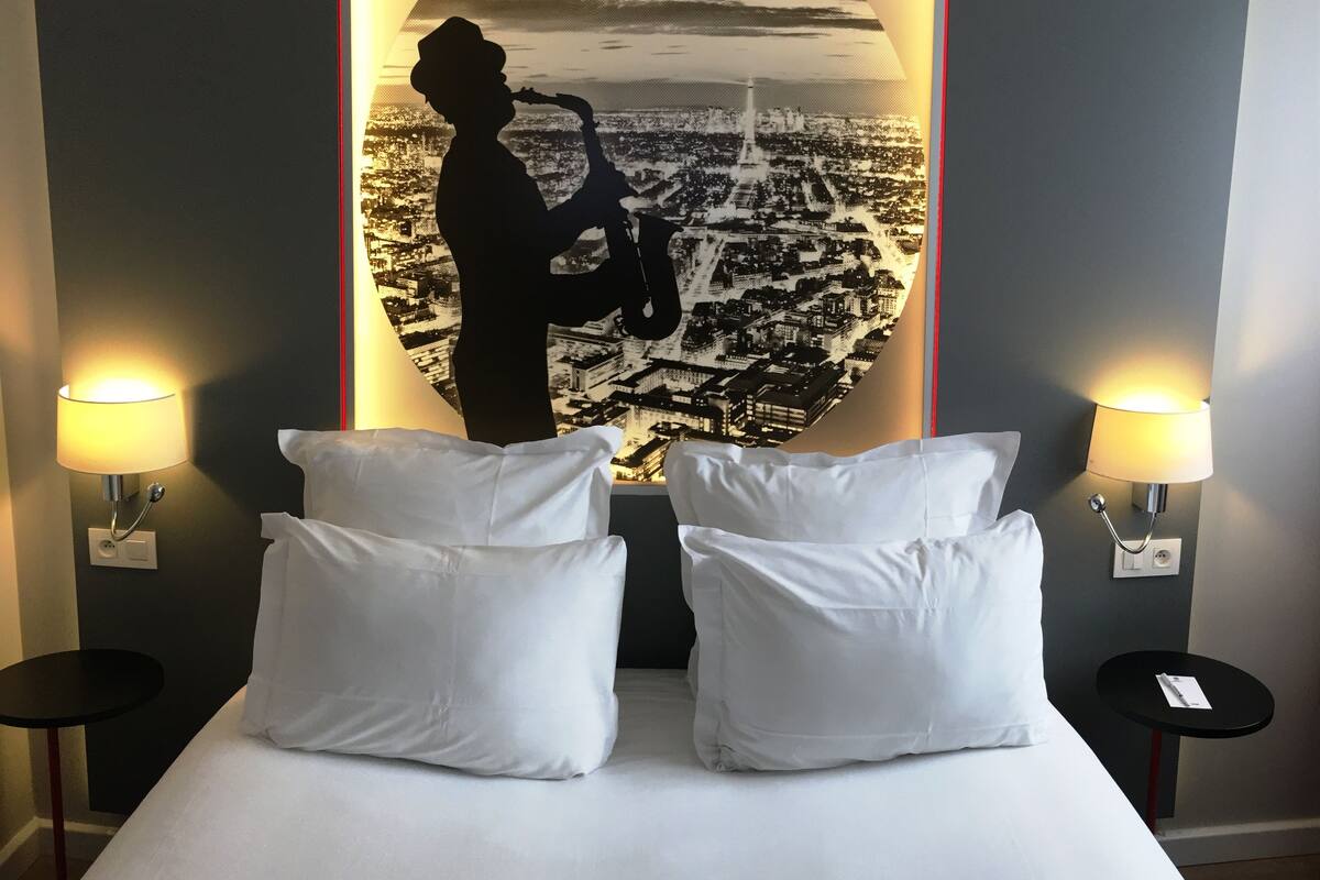 Habitación de hotel con cama, dos almohadas blancas, dos mesitas de noche con lámparas y un mural grande con un paisaje urbano.