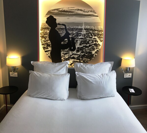 Une chambre d’hôtel avec un lit, deux oreillers blancs, deux tables de chevet avec des lampes et une grande fresque murale représentant un paysage urbain.