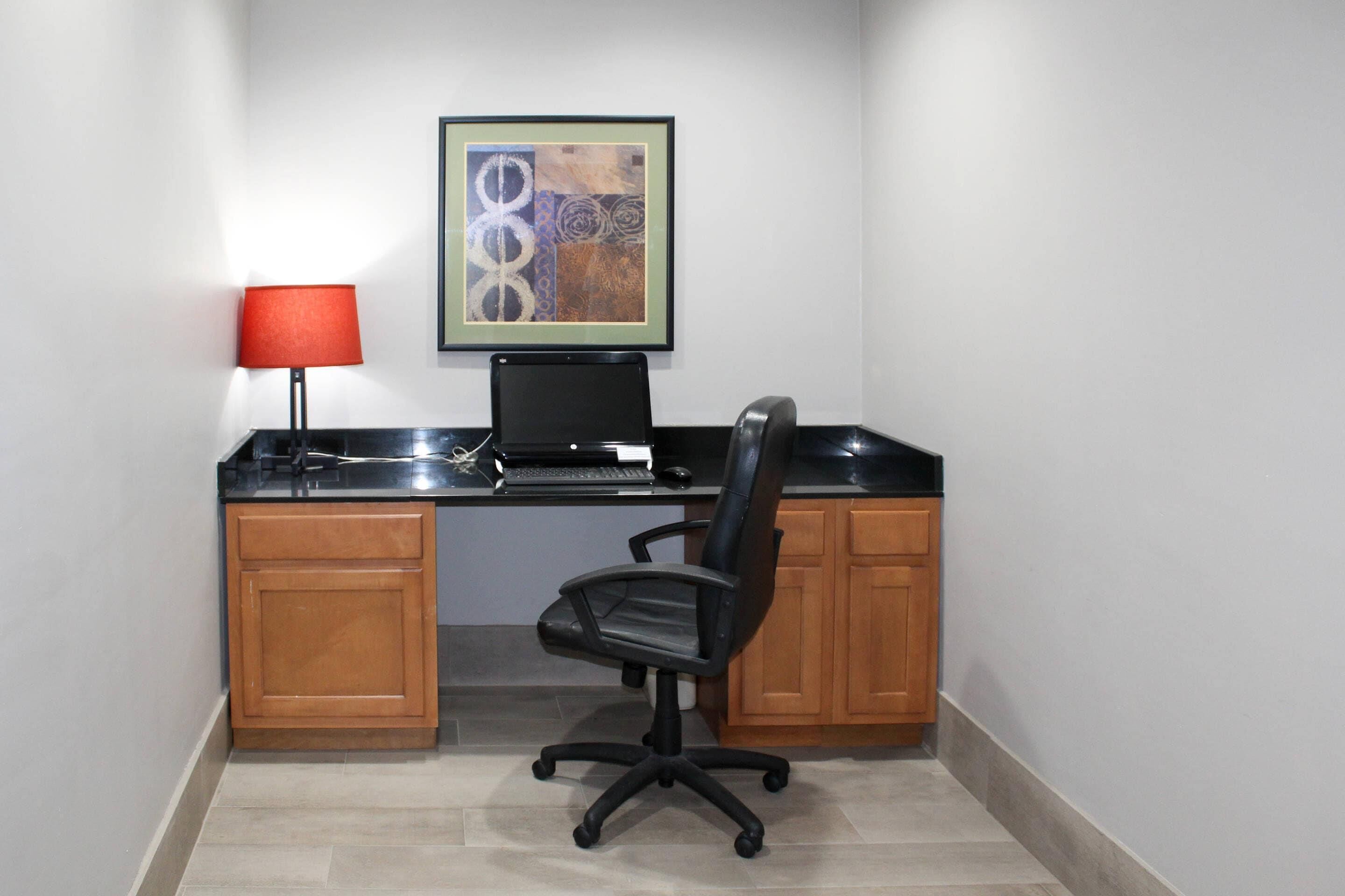 A small office space with a desk, a chair, a computer, a red lamp, and a framed artwork on the wall.