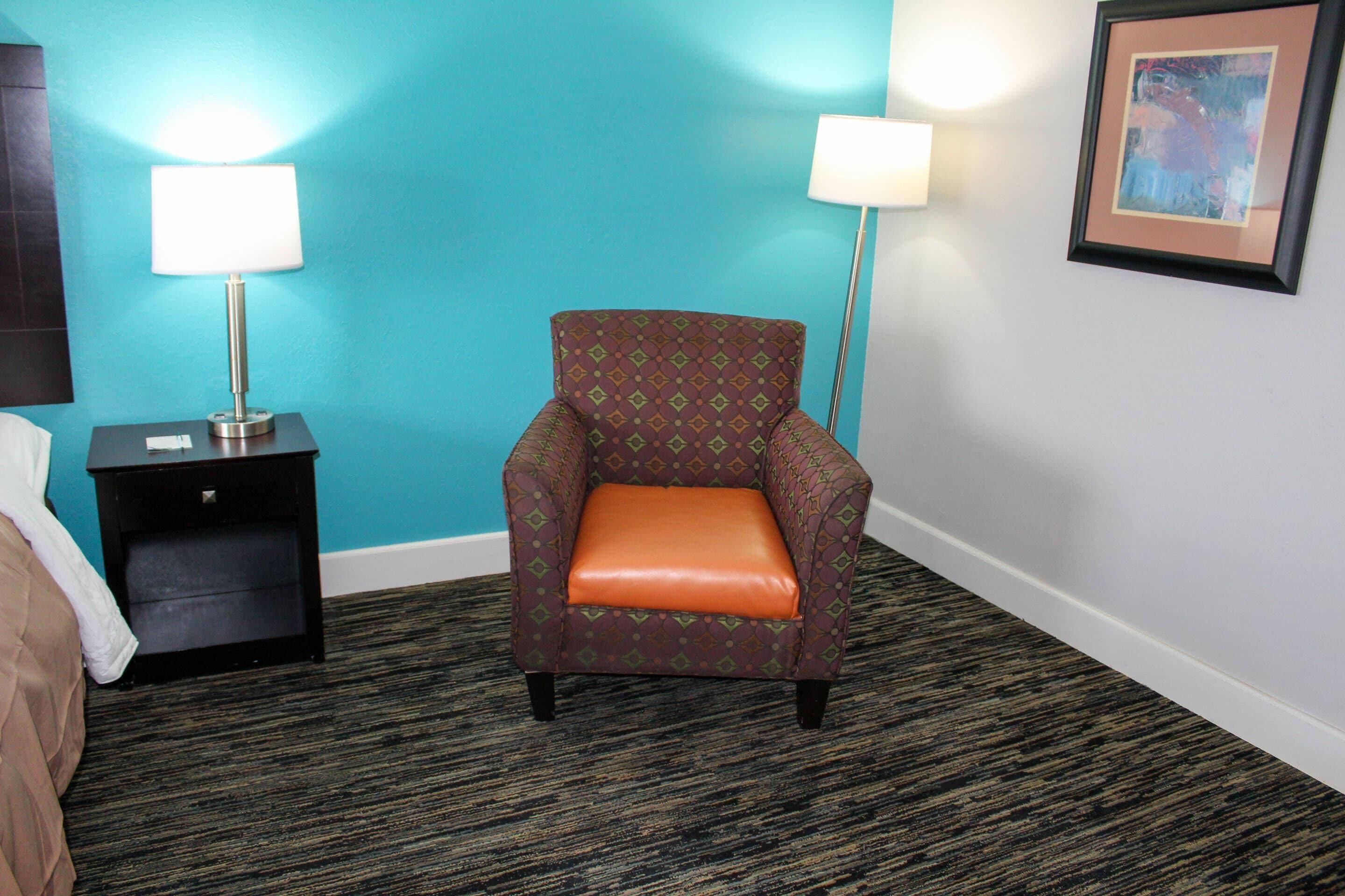 Une chambre d’hôtel avec un mur de couleur turquoise, un fauteuil à motifs agrémenté d’un coussin orange, une petite table de chevet avec une lampe et un tableau encadré accroché au mur.