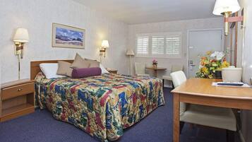 A hotel room with a bed, bedside tables, a desk, a chair, a window with blinds, and a wall-mounted lamp.