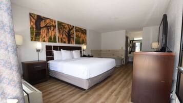 A hotel room with a large bed, a wooden floor, a desk, a chair, a nightstand, a mirror, and a television.