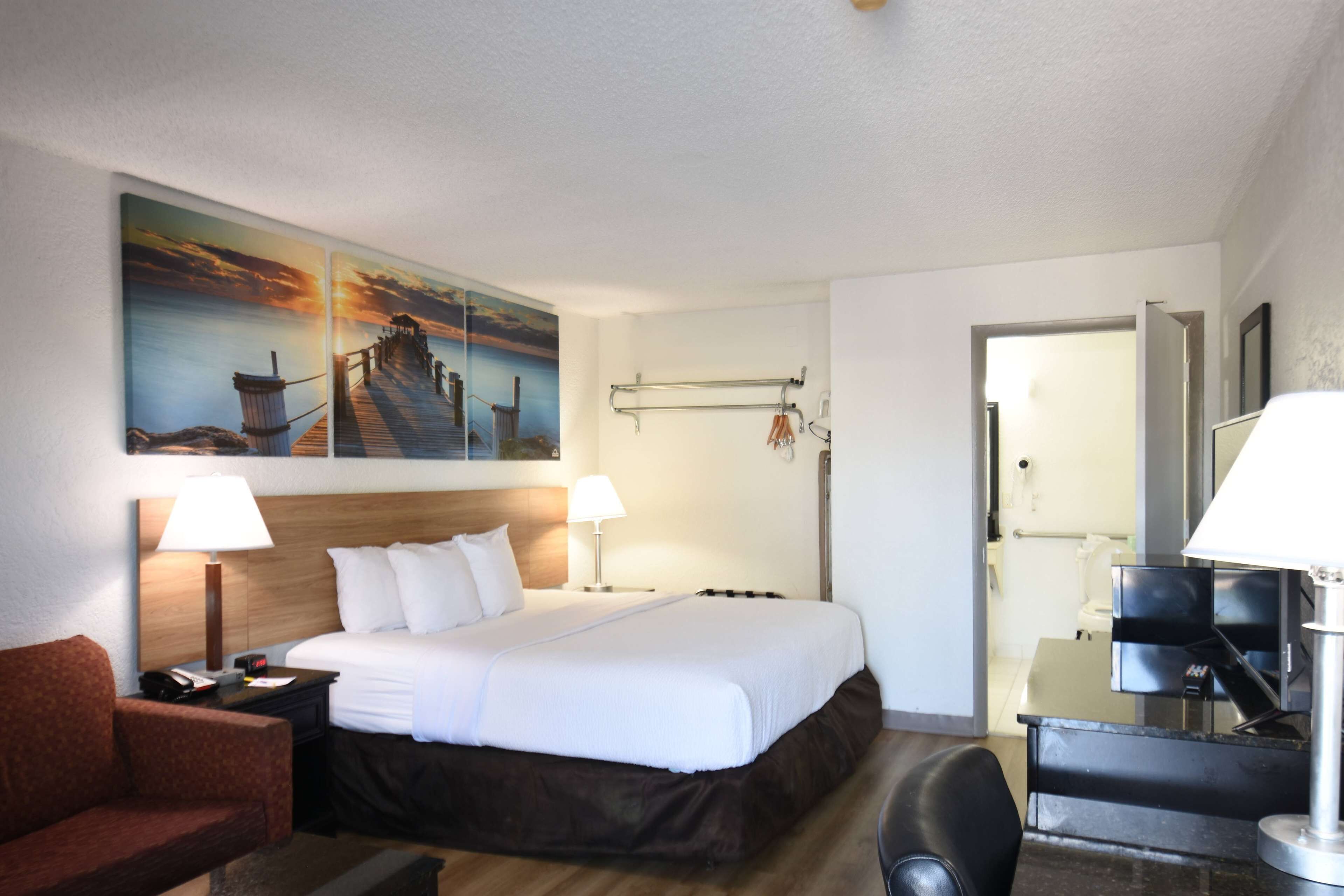 Une chambre d’hôtel avec un grand lit, deux lampes de chevet, une tête de lit en bois, un tableau représentant un coucher de soleil, un bureau avec une chaise et un miroir.