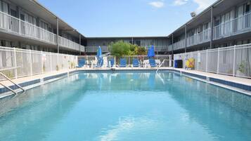 A swimming pool with blue chairs and umbrellas, surrounded by a white fence, with a building in the background.
