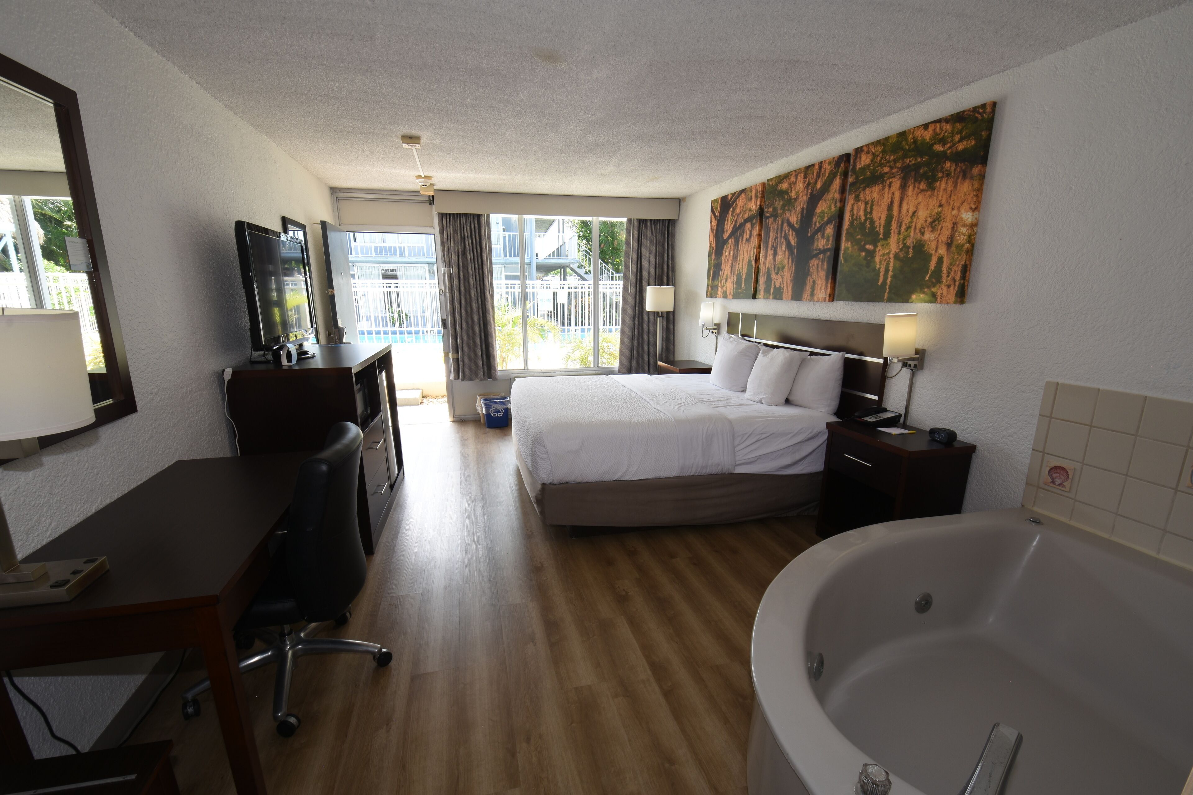 Habitación de hotel con cama, escritorio, silla, lámpara de escritorio, televisor, ventana con cortinas, un cuadro y bañera.