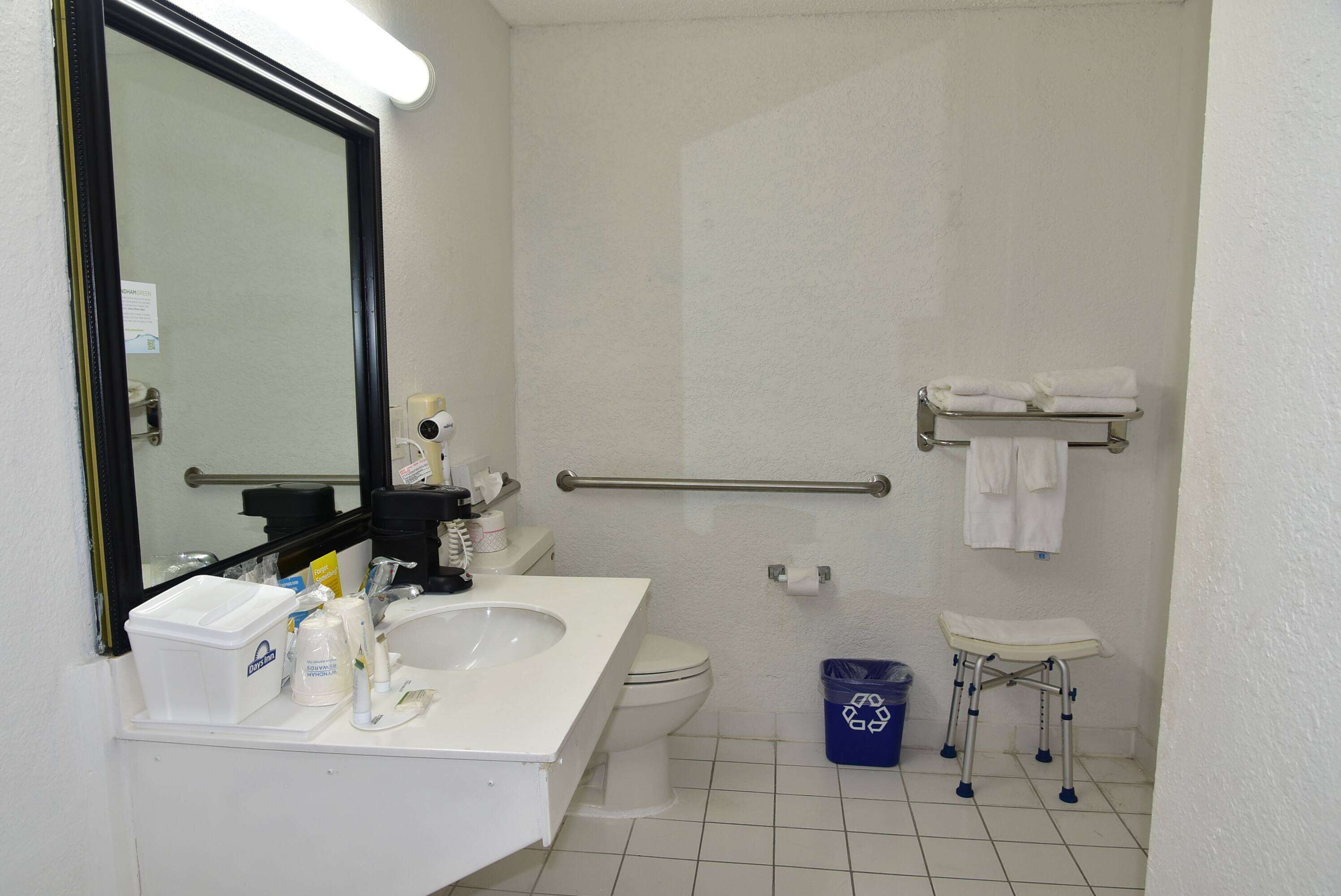 Une salle de bain avec un lavabo, des toilettes et une douche, le tout de couleur blanche. On y trouve un miroir, un porte-serviettes et une barre d’appui.