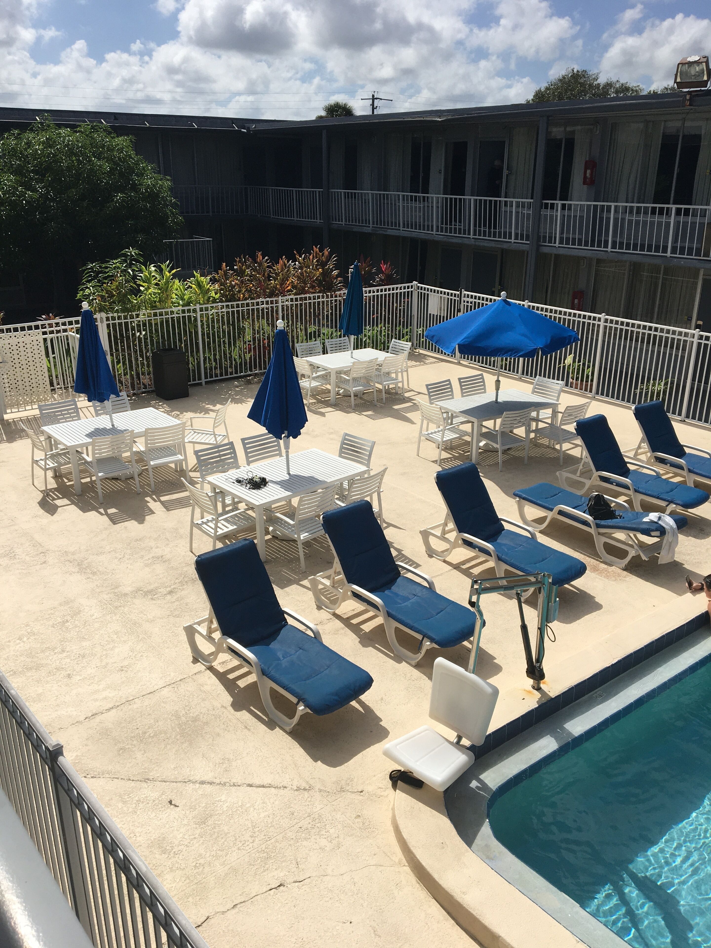 A poolside area with white lounge chairs and blue umbrellas, surrounded by a white fence and a building in the background.