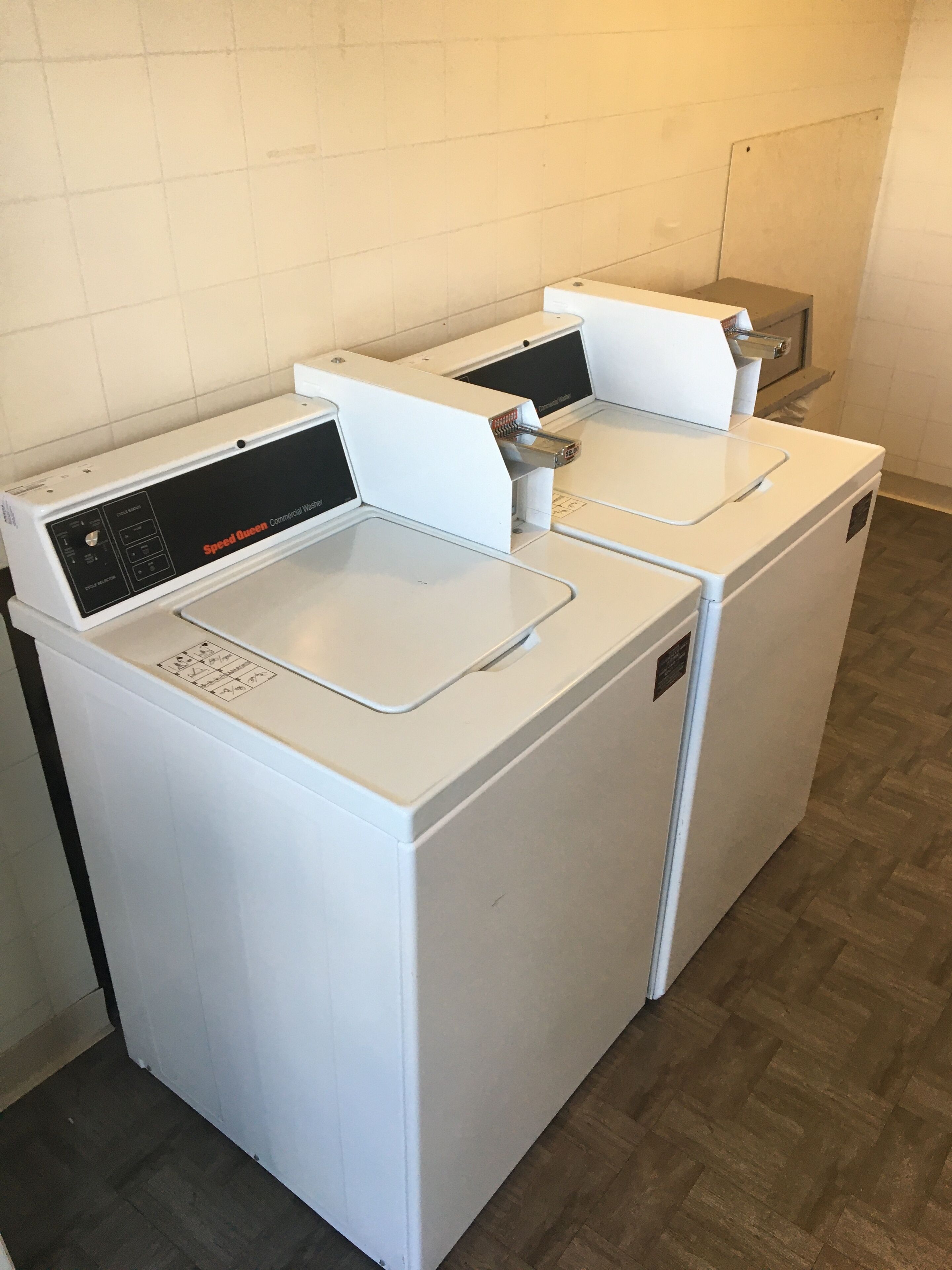 A row of white commercial washers with control panels, in a room with wooden flooring.