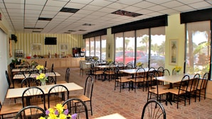 A restaurant with multiple tables and chairs, a counter with a coffee machine, and large windows with a view of parked cars and palm trees.